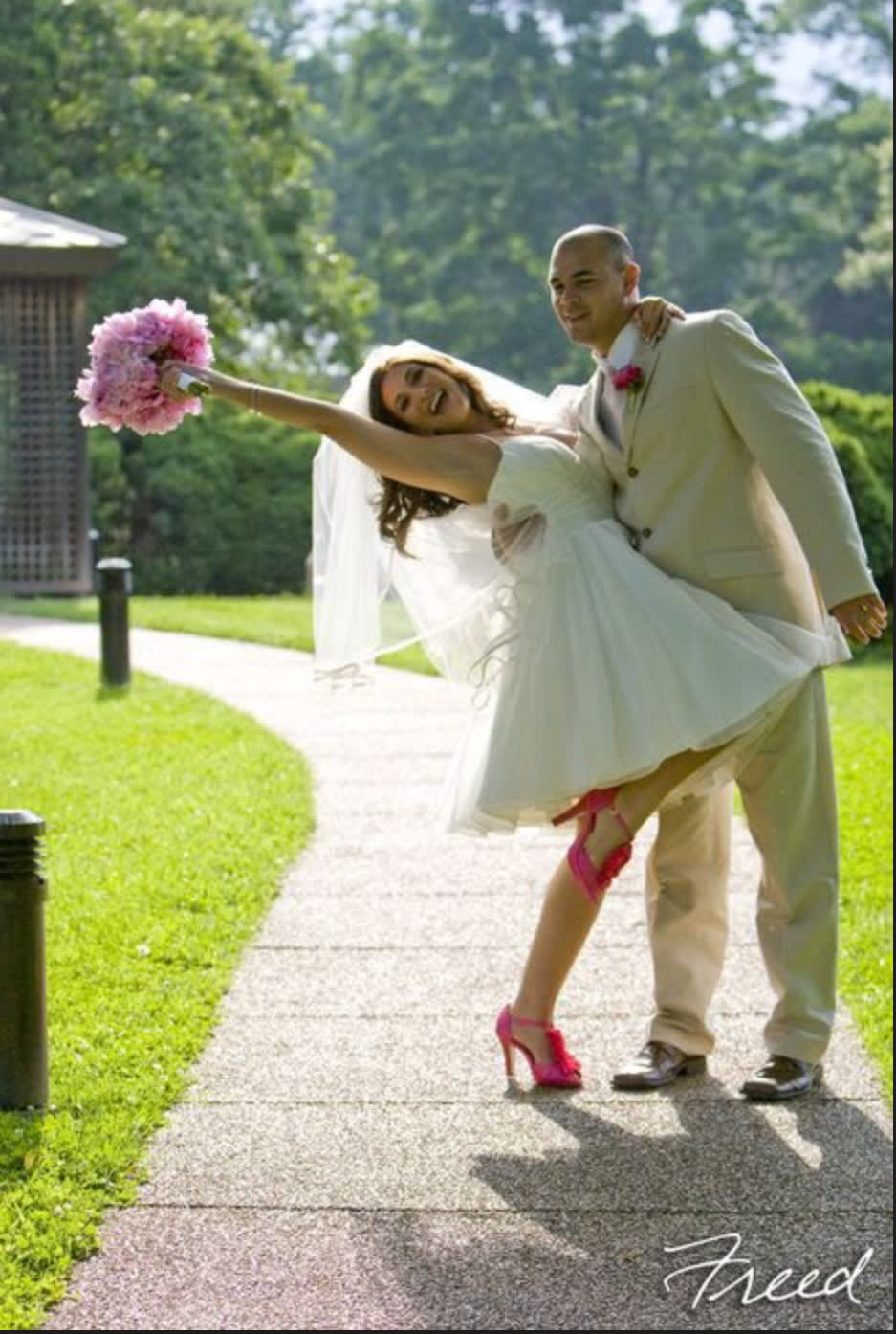 Bride being carried by groom, holding a pink bouquet outdoors