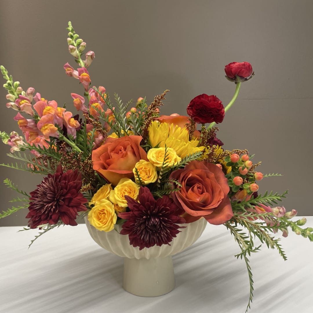 Mixed bouquet in a white pedestal vase with orange, yellow, and burgundy flowers