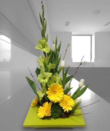 Tall floral arrangement with yellow gerbera daisies and green lilies on a square base