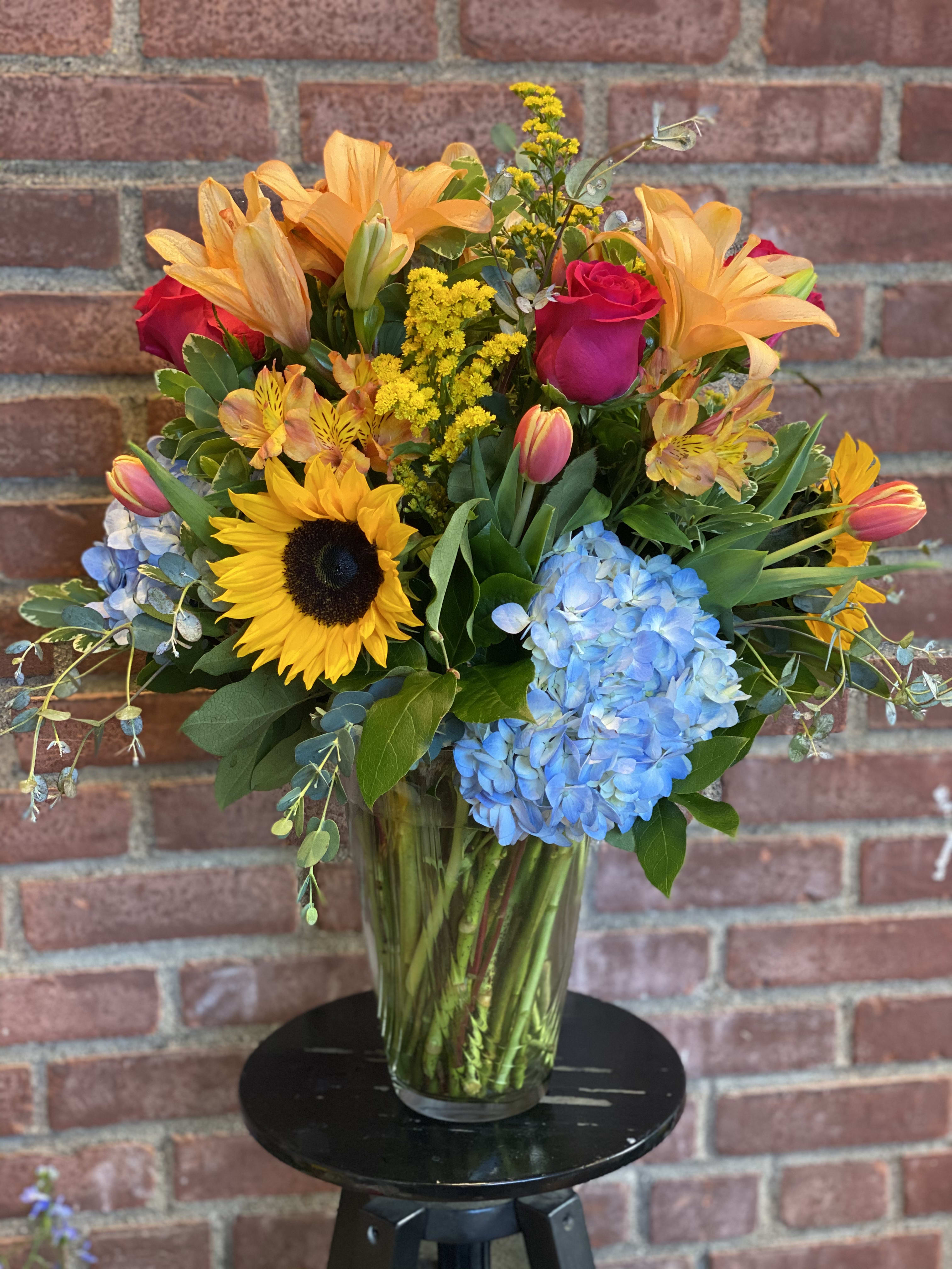 Mixed bouquet with sunflowers, roses, lilies, and blue hydrangeas in a glass vase
