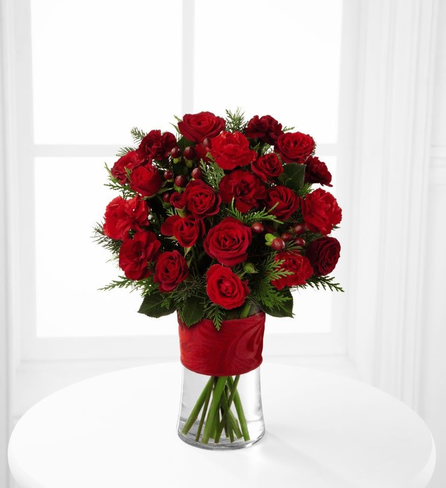 FTD® Spirit of the Season™ Bouquet - The FTD® Spirit of the Season™ Bouquet bursts with beauty and cheer to dazzle your special recipient with its holiday beauty. Brilliant red spray roses and mini carnations are brought together with burgundy mini carnations, red hypericum berries and assorted holiday greens in a clear glass vase accented with a thick red designer ribbon to create a special sentiment of seasonal wishes and caring kindness. 