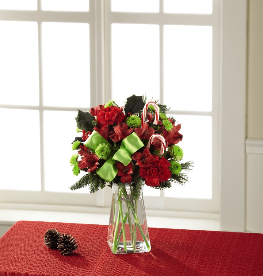 The FTD Candy Cane Lane Bouquet - Stacking the odds of holiday fun in your recipient's favor, this fresh flower bouquet is a Christmas favorite that never goes out of style. Frilly red carnations are paired with red Peruvian Lilies surrounded by green button poms, variegated holly, and an assortment of fragrant Christmas greens. Accented with a bright green satin ribbon and classic candy canes, this festive holiday bouquet is that perfect way to say, "Merry Christmas," to the ones you love throughout the yuletide season.