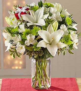 FTD Wrapped Up In Joy Holiday Bouquet - white Asiatic Lilies, white Peruvian Lilies, green button poms, assorted holiday greens with a red present pick and a clear glass vase. Bouquet is approximately 14"H x 13"W.
