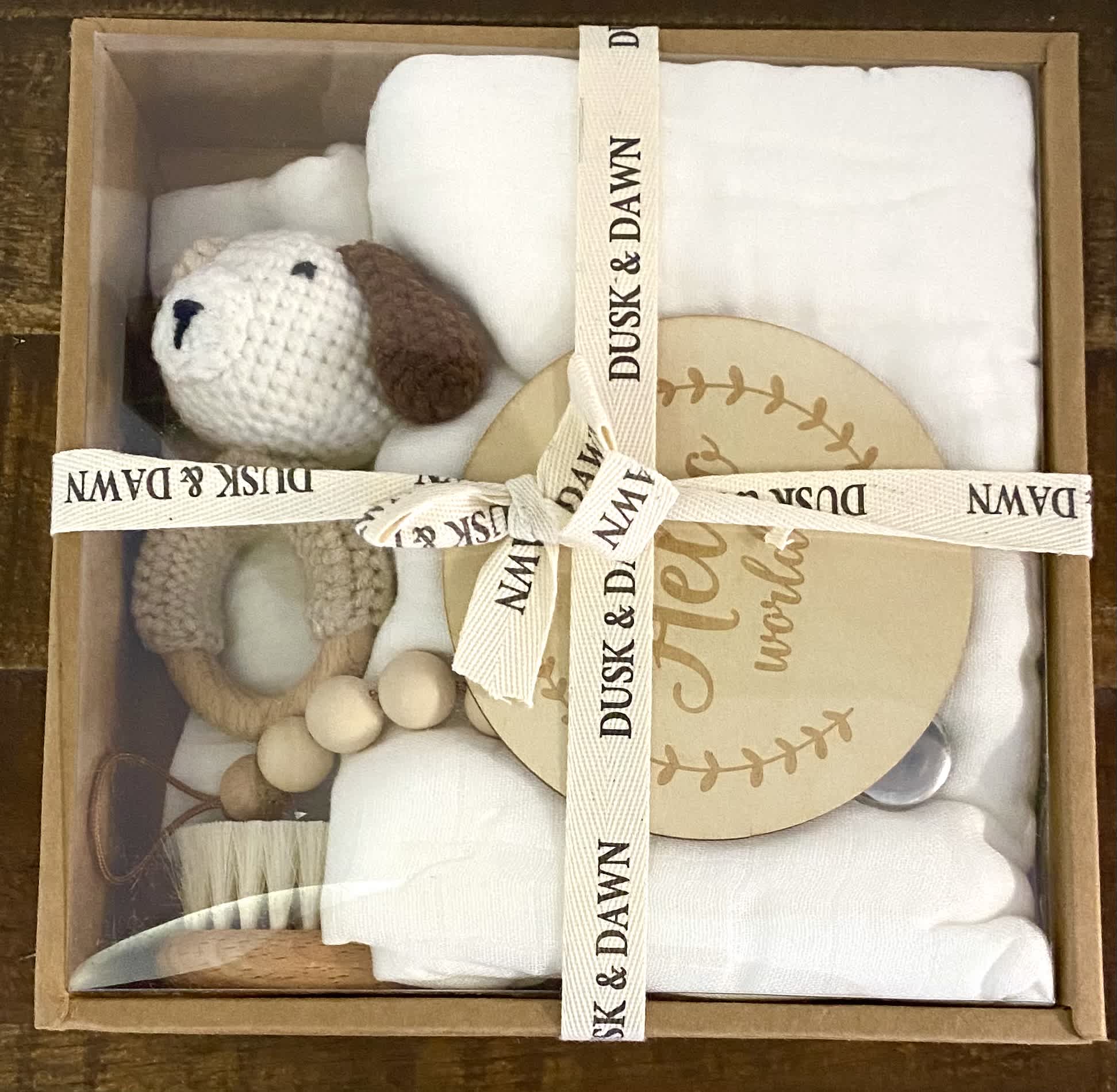 Welcome Baby Gift Set  - charming 6-piece Baby Gift Set, thoughtfully curated to delight both parents and their precious little ones. This ensemble includes everything a baby needs, beautifully presented in a rustic kraft box adorned with a clear lid and tied with a soft cotton ribbon.  Inside, you'll discover a collection of essentials designed for comfort and style, making it the perfect gift for baby showers or welcoming a new arrival.  Each Gift Set Includes:  One Pacifier Clip One Bamboo Baby Hairbrush One Hello World Baby Announcement Sign One Bunny Rattle One Muslin Bandana Bib One Muslin Organic Bamboo Swaddle
