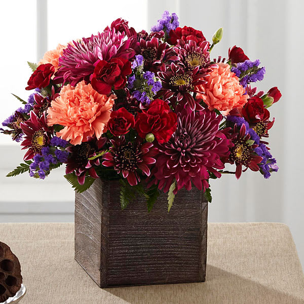 The Homespun Harvest Bouquet - A nod to the rich deep colors of the autumn season this inspiring bouquet adds a touch of glamour to the harvest months ahead. Burgundy chrysanthemums plum chrysanthemums red mini carnations and purple statice are highlighted by pops of orange carnations all while situated in a rustic square wooden container. An excellent way to send a touch of fall beauty to your recipient to help them bring the magic of the season into their home. GOOD bouquet includes 11 stems. Approx. 10"H x 10"W. BETTER bouquet includes 15 stems. Approx. 11"H x 12"W. BEST bouquet includes 19 stems. Approx. 12"H x 13"W.