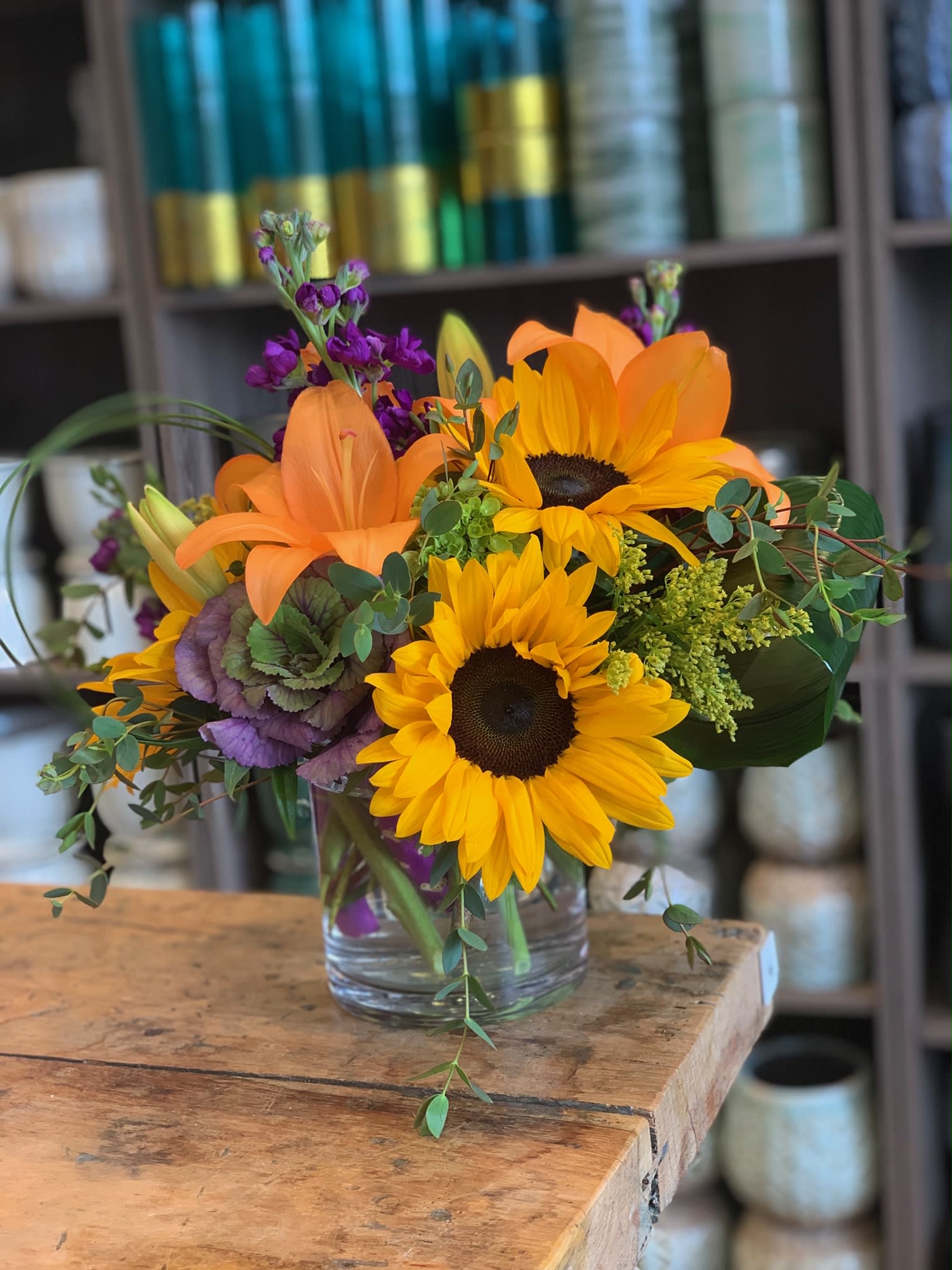 Ray of sunshine - Designed in a sleek cylinder vase, this bold and textural arrangement features cheerful sunflowers, striking ornamental kale, vivid orange lilies, fragrant stock, and whimsical loops of bear grass. A perfect blend of rustic charm and modern flair, ideal for brightening any space.  