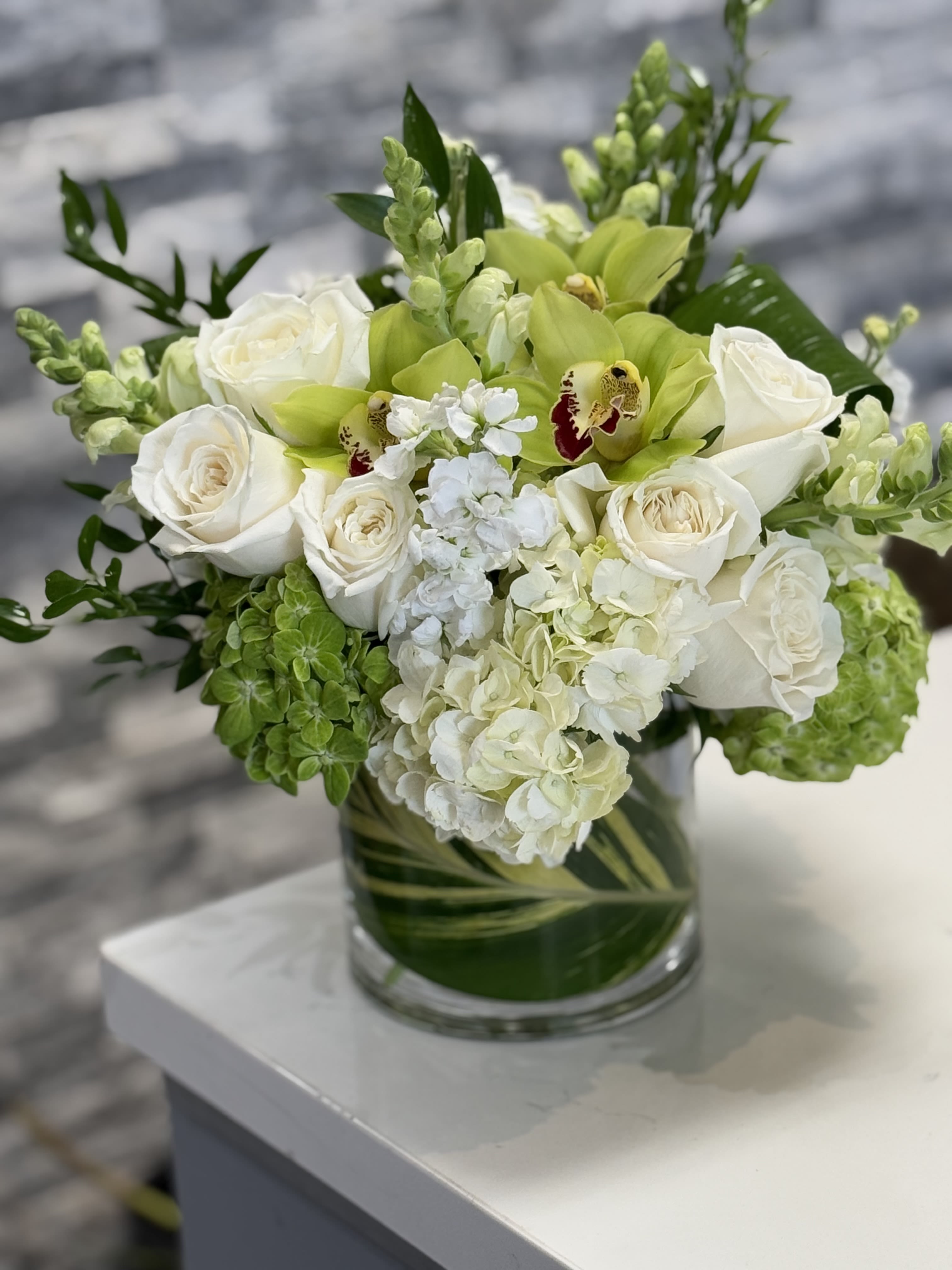 Daydream  - A beautiful floral arrangement in a clear glass vase, styled in a modern, elegant design. White Roses, Green Cymbidium Orchids, White Hydrangeas, Green Hydrangea,  White Stock, White Snapdragons, and assortment of mixed greens. In a clear cylinder vase.