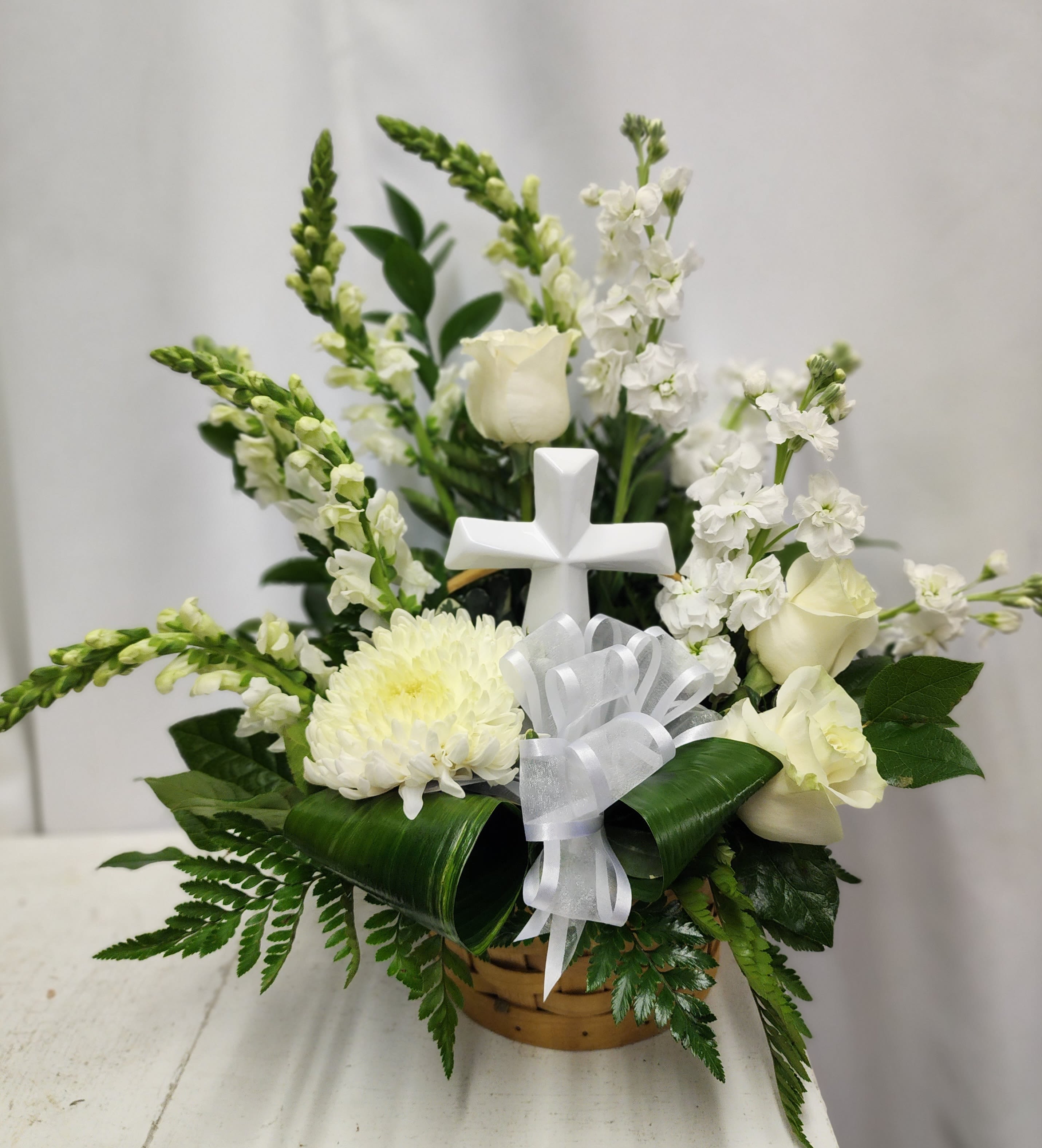 Basket of Glory - An all white basket of flowers with a keepsake white modern cross, created in a honey color basket with white bow. Includes snapdragons, stock, roses and cremone. Simple elegant and thoughtful for any sympathy service.