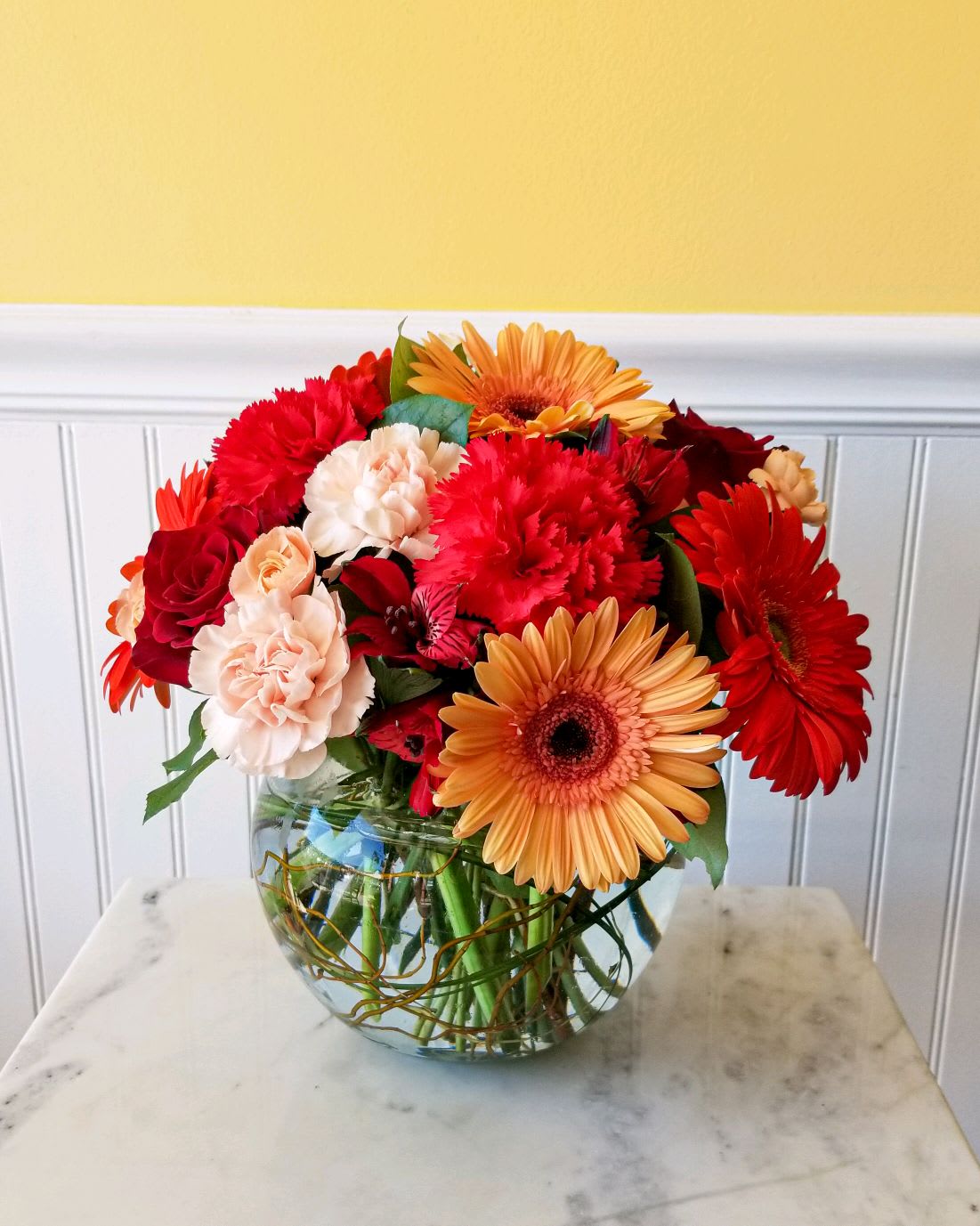 Mixed red, peach, and orange flowers arranged in a glass bowl vase