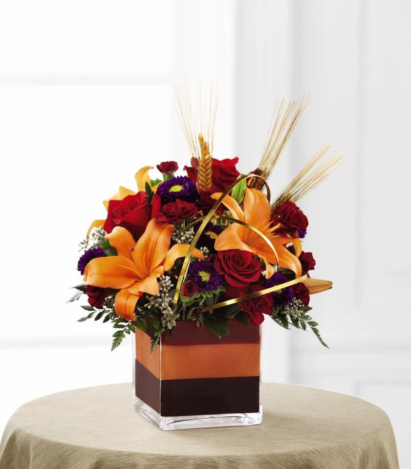 The FTD Autumn Passages Bouquet - The FTD Autumn Passages Bouquet is blooming with rich autumn colors to offer your special recipient a warm wish for a wonderful fall season. Brilliant red roses, orange Asiatic lilies, burgundy mini carnations, purple matsumoto asters, dried wheat stems, and a variety of lush greens are beautifully arranged in a clear glass cubed vase featuring layers of colored foam inside for an incredible look. Elegantly accented with gold flat wire throughout the arrangement, this bouquet creates the perfect seasonal offering. 