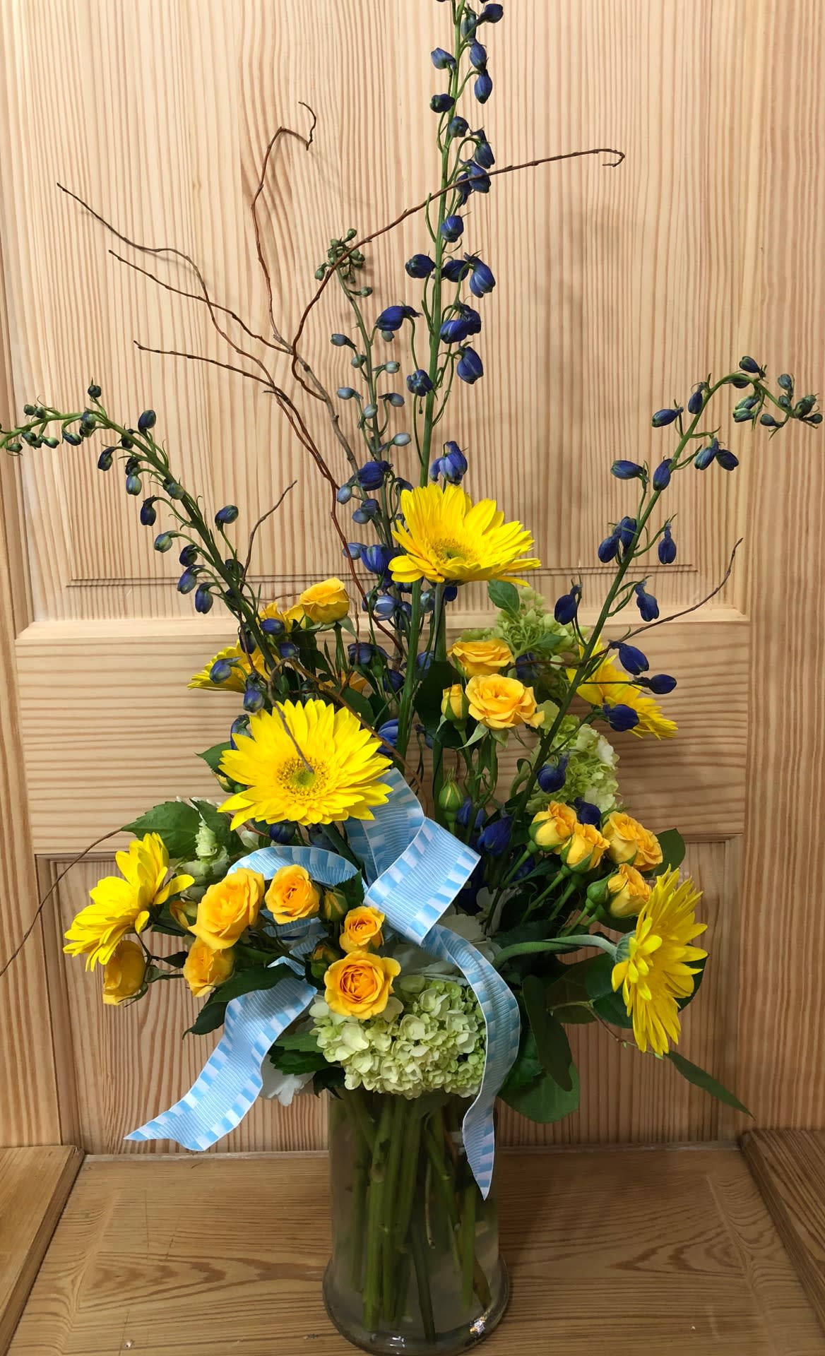 Yellow gerberas and roses in a glass vase with blue ribbon accents