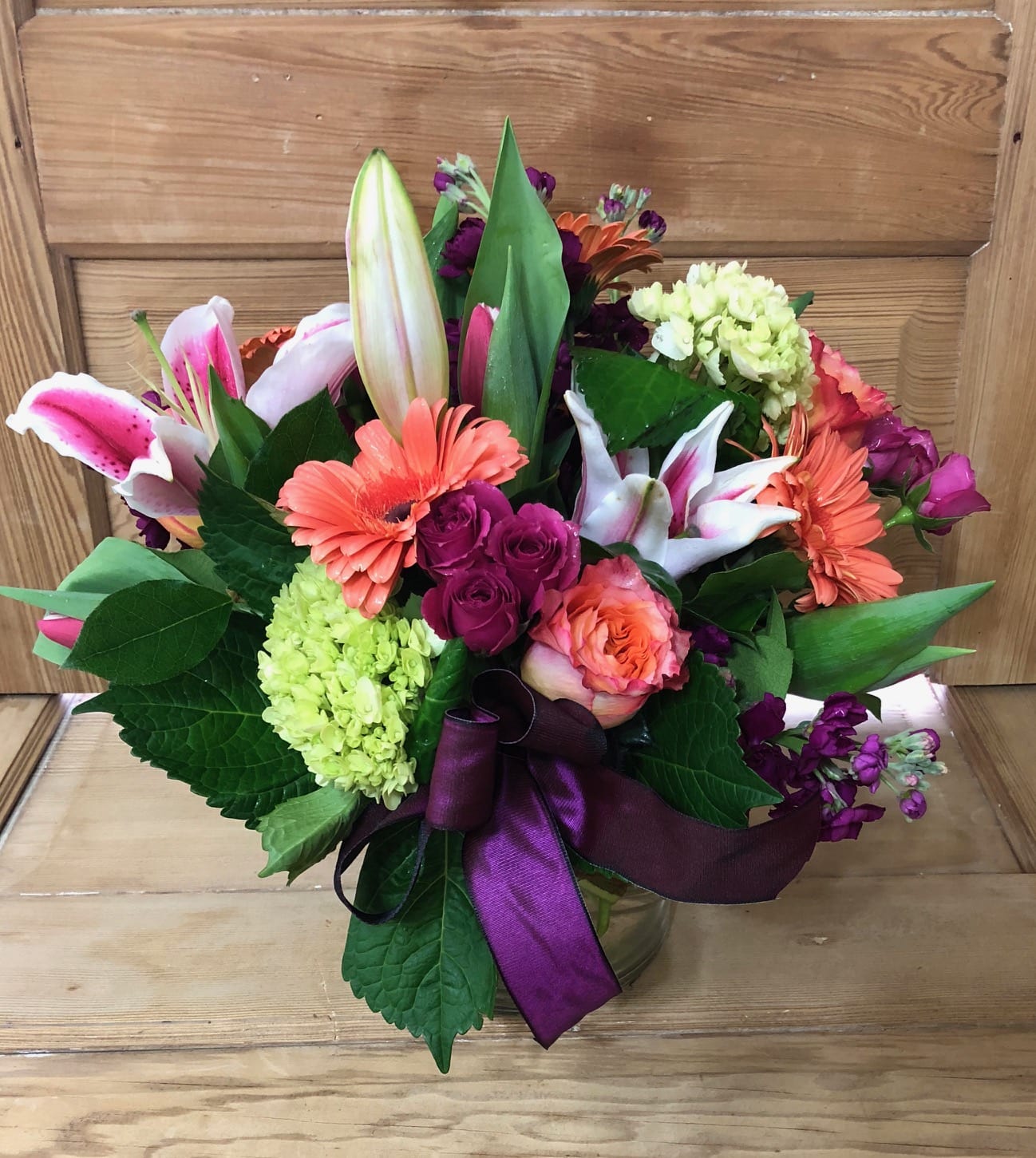 Mixed bouquet of lilies, roses, and gerbera daisies in a glass vase with a purple ribbon