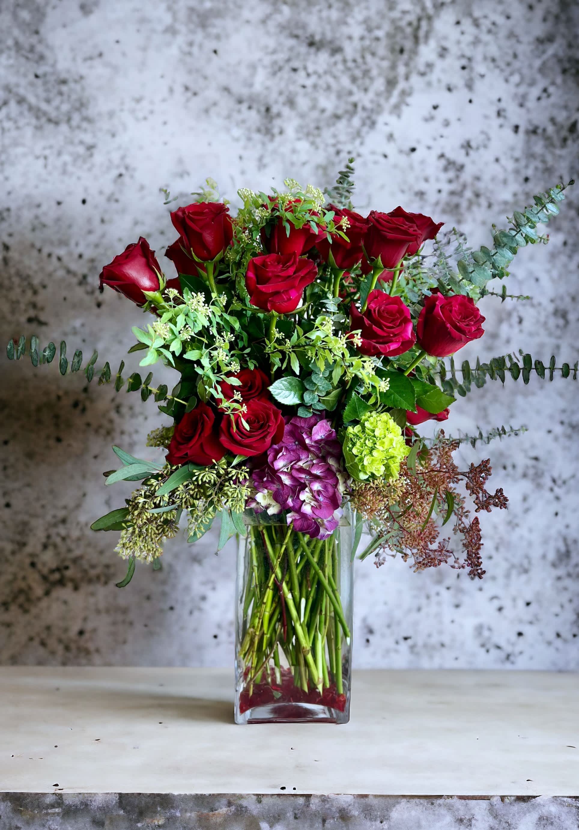 Red roses arranged in a clear glass vase with mixed greenery and purple hydrangea