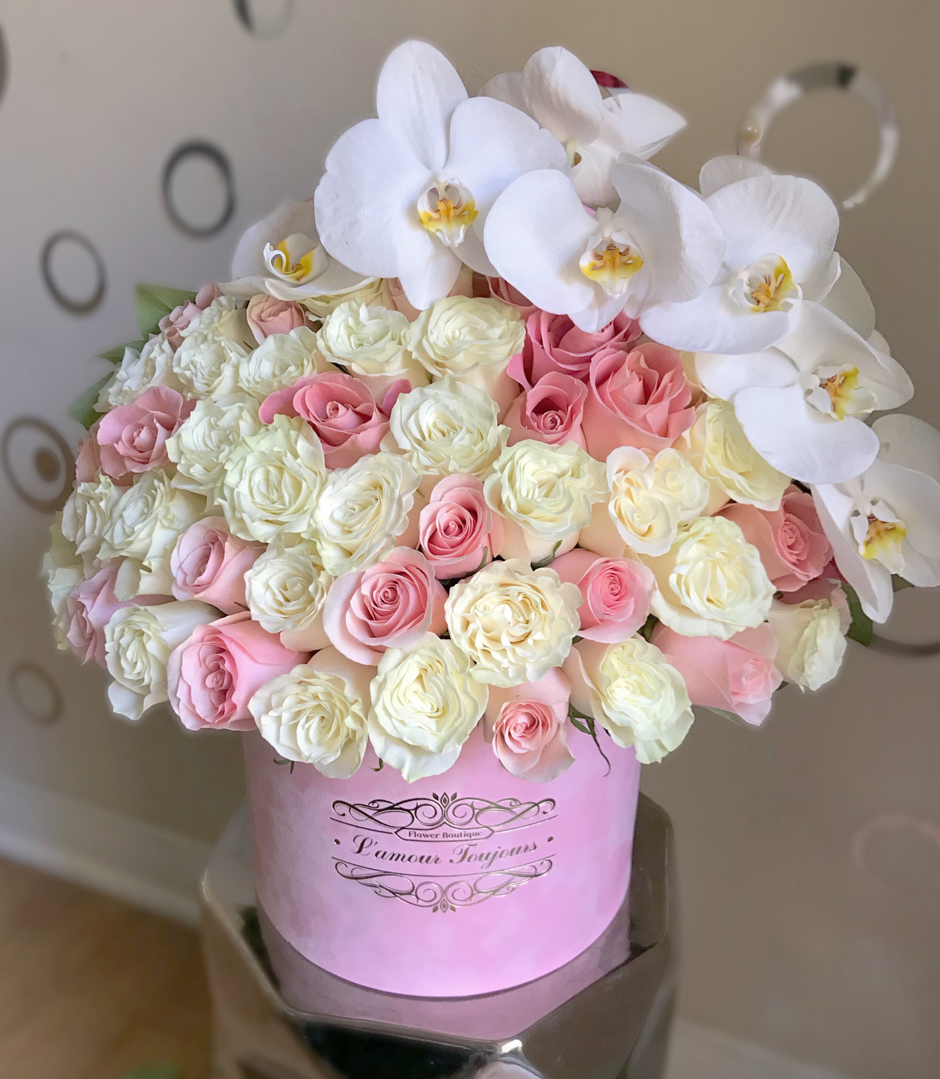 Round box arrangement of white orchids and pink and cream roses in a pink hatbox.