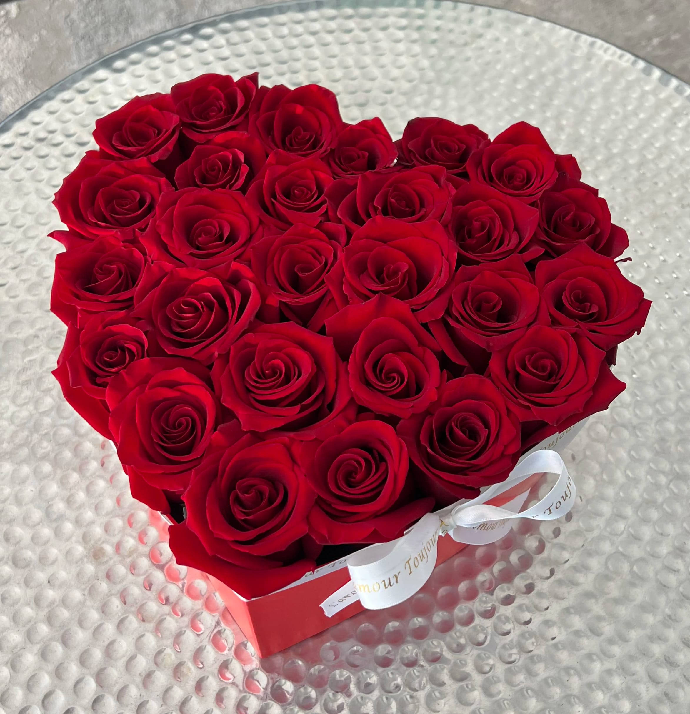 Heart-shaped box filled with red roses and tied with a white ribbon on a glass table