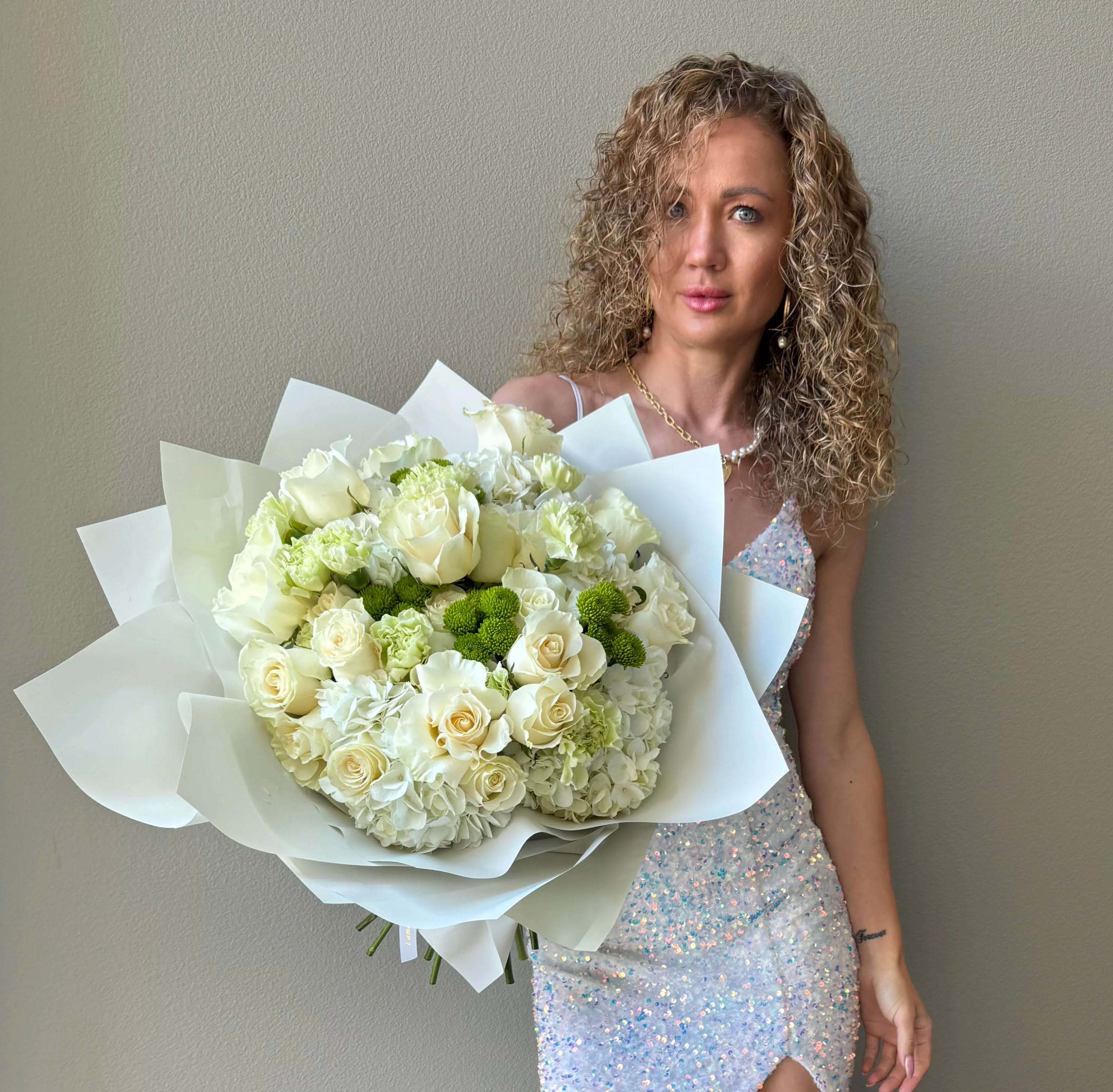 Large white and green bouquet of roses and hydrangeas wrapped in paper, held by a woman in a sequin dress