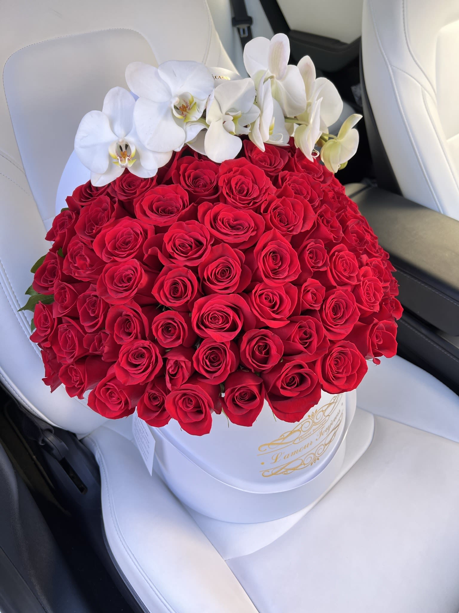 100 Roses + stem of orchids - A dramatic dome of 100 velvety red roses arranged in a crisp white round hatbox creates an unforgettable anniversary or Valentine's Day statement - perfect for Mother's Day, a milestone birthday, or any moment when you want someone to feel deeply cherished. A graceful arc of large white Phalaenopsis orchid blooms traces the top edge, adding sculptural contrast and an elegant, modern silhouette that reads luxe and romantic at first glance.  The arrangement is presented in a white round hatbox with subtle gold script, echoing the orchids' clean white tones and giving the design a polished, gift-ready finish. Rich red roses are tightly packed to form a full, compact dome while a single stem of white phalaenopsis orchids curves across the surface for texture and visual lift. Ideal for celebrations from anniversaries and weddings to graduations, housewarmings, and get-well wishes, this striking red-and-white bouquet also suits holiday moments like Christmas, Mother's Day, or an Easter centerpiece.  For care, keep the box in a cool spot away from direct sunlight and drafts; refresh any visible water source according to the hatbox style to maintain bloom life. This is the kind of arrangement that makes the recipient feel truly seen - a bold, romantic gesture for love and celebration or a show-stopping surprise delivered "just because."