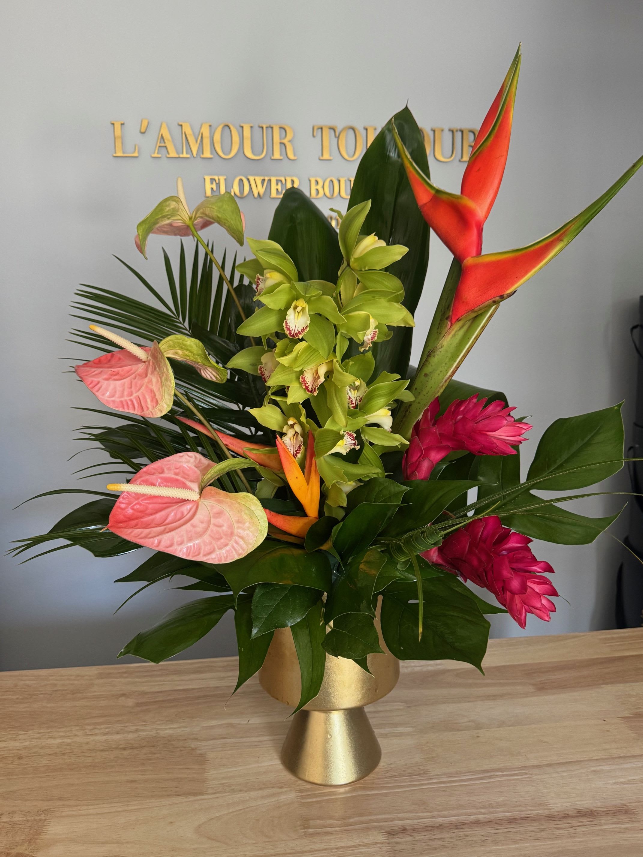 Tropical arrangement with green orchids, pink blooms, and orange heliconia in a gold pedestal vase