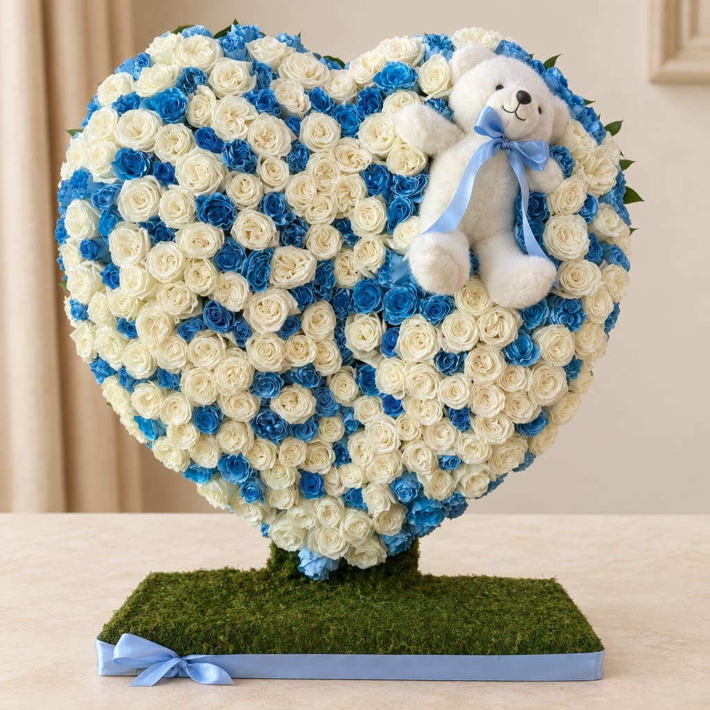 Heart-shaped rose arrangement with a teddy bear and blue ribbon on a grass-covered base