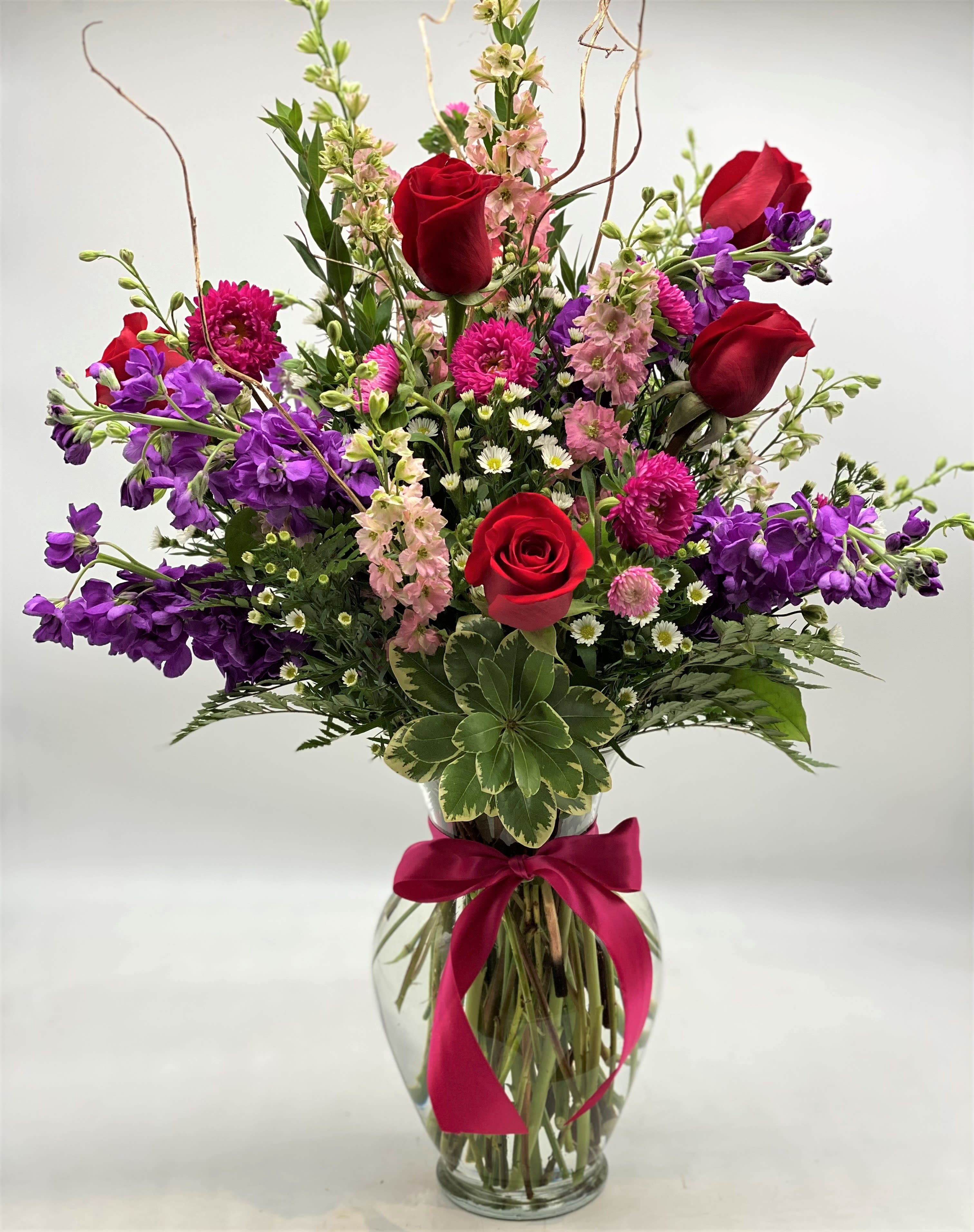 Mixed bouquet of red roses, purple blooms, and pink flowers in a glass vase with ribbon