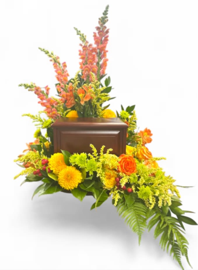 Summer Memories Memorial Urn (Wood Box)  - This vibrant sympathy arrangement, thoughtfully presented in an 8-inch design tray, offers a heartfelt tribute filled with warmth and beauty. A foundation of lush foliage, including variegated pittosporum, sword fern, ming fern, eucalyptus, and lily grass, creates a harmonious backdrop that enhances the floral display. Four cheerful yellow snapdragons stand tall, symbolizing strength and grace, while four lively stems of green button poms add a playful touch. Three stems of orange spray roses bring bursts of color, representing admiration and joy, complemented by two stems of bright yellow alstroemeria, which express friendship and devotion. The addition of four solidago stems adds a touch of golden warmth, alongside four red hypericum stems, symbolizing protection and encouragement. Striking green spider mums offer a unique elegance, and three bi-colored orange roses complete the arrangement, embodying a vibrant spirit and the beauty of cherished memories. Together, these flowers convey a message of love, comfort, and remembrance during this poignant time. Urn not included.