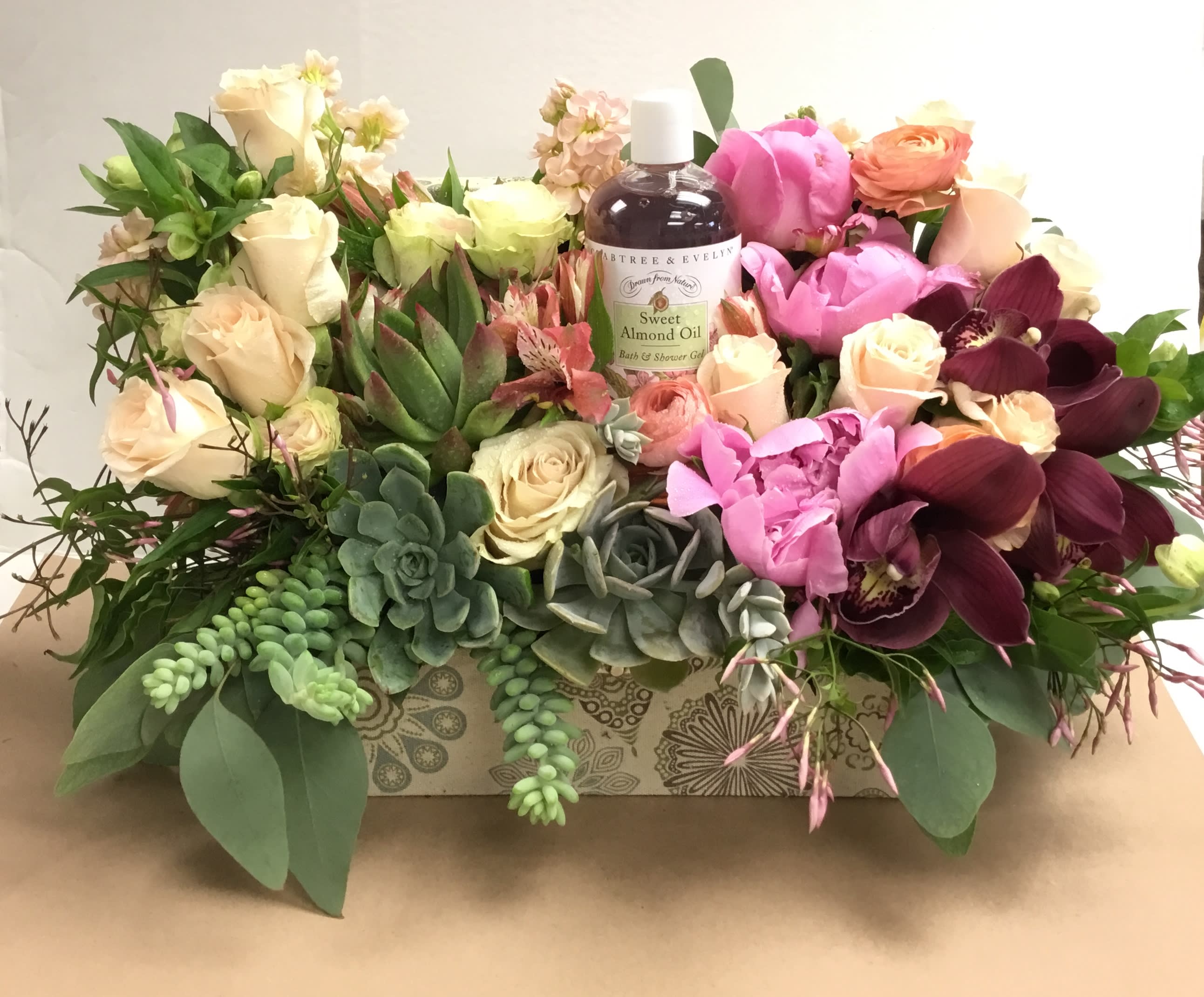 Mixed floral arrangement with roses, orchids, succulents, and a bath oil bottle in a decorative box
