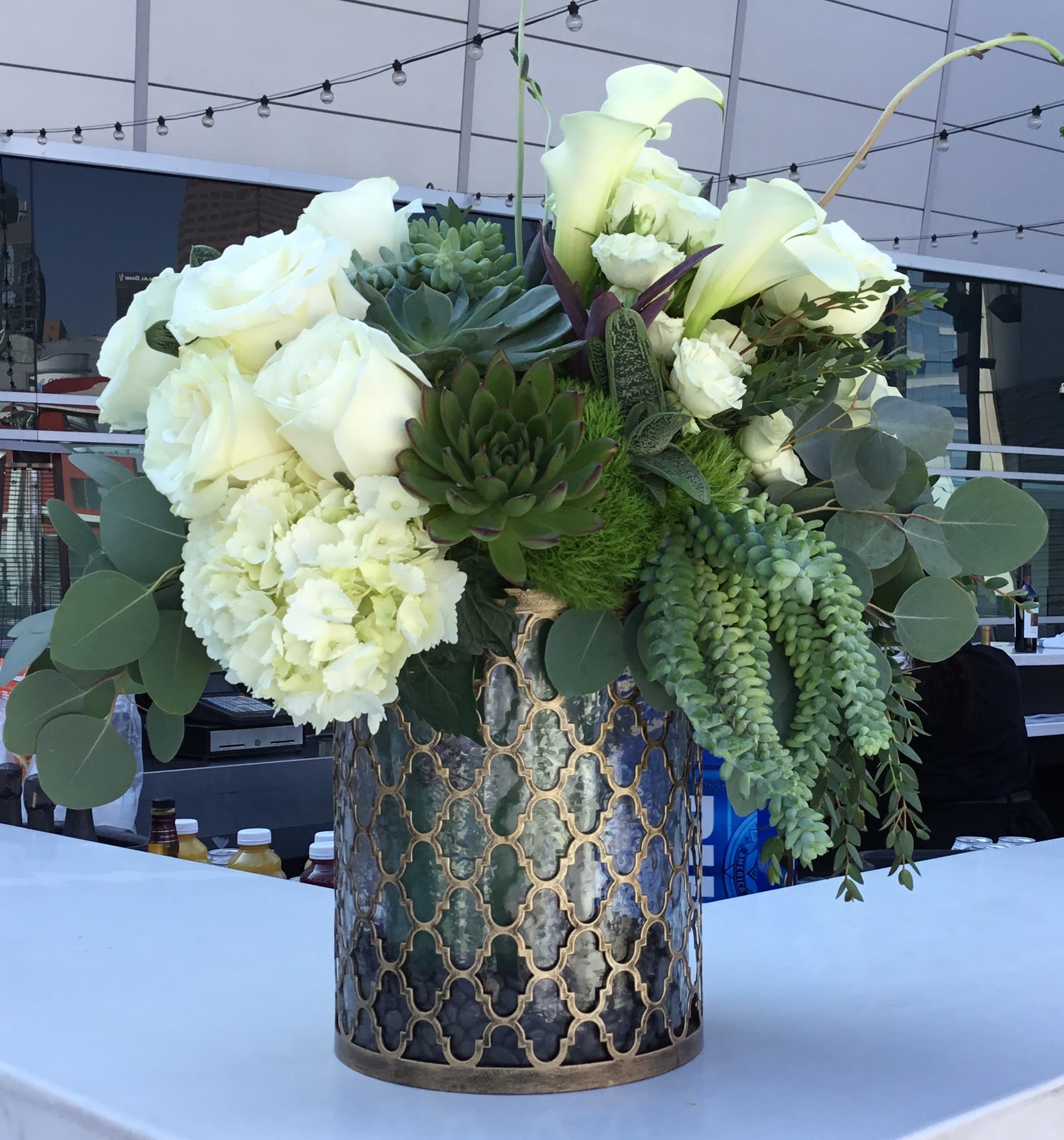 White roses and calla lilies arranged with succulents in a patterned metal vase