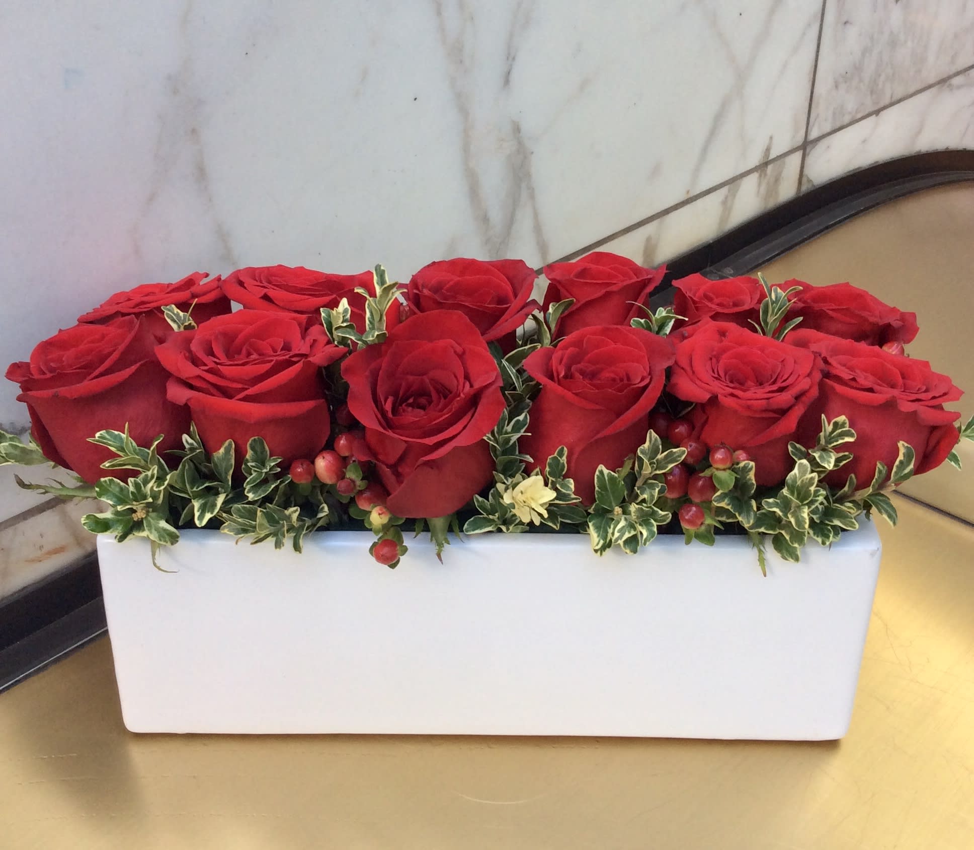 Red roses arranged in a white rectangular vase with small variegated greenery