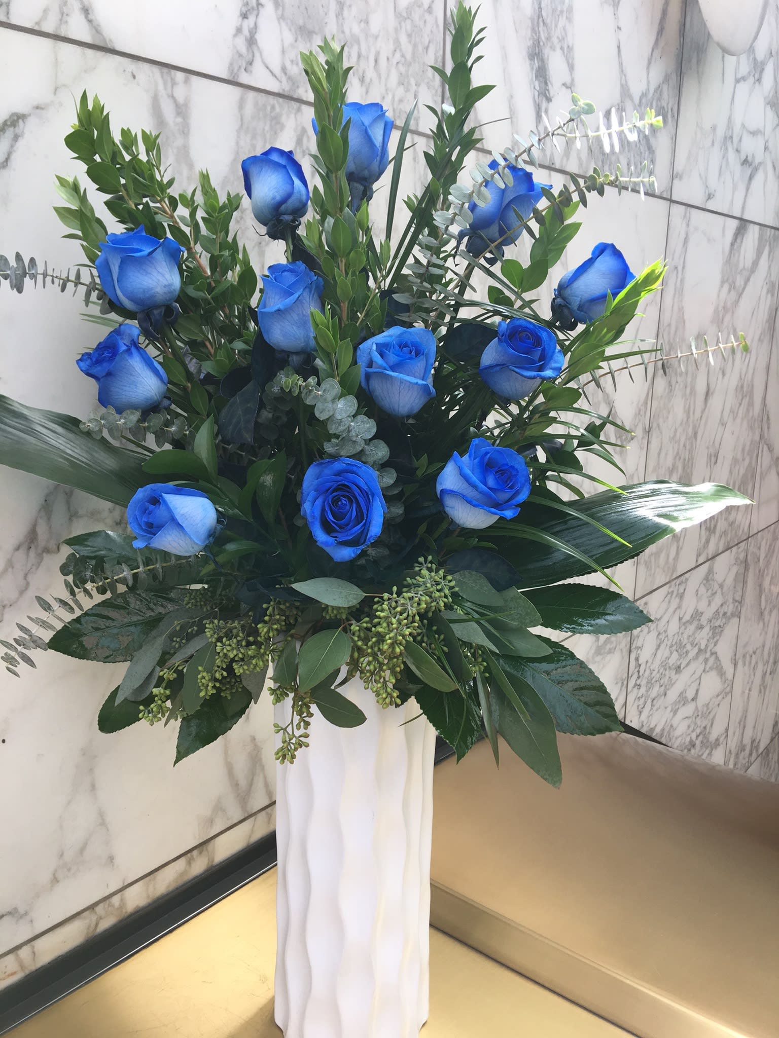 Blue roses in a tall white vase with mixed greenery