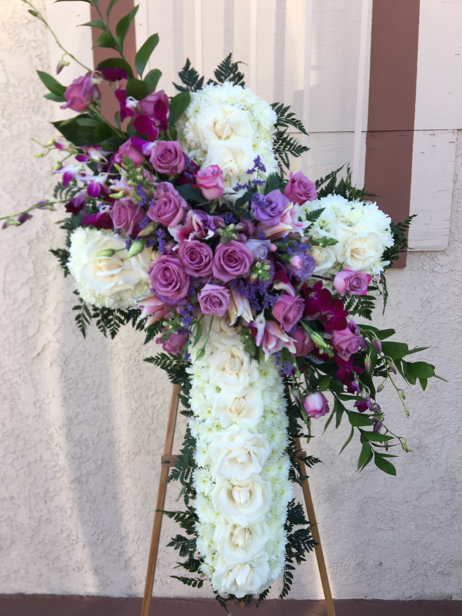 Cross-shaped floral tribute with white roses and purple flowers on an easel