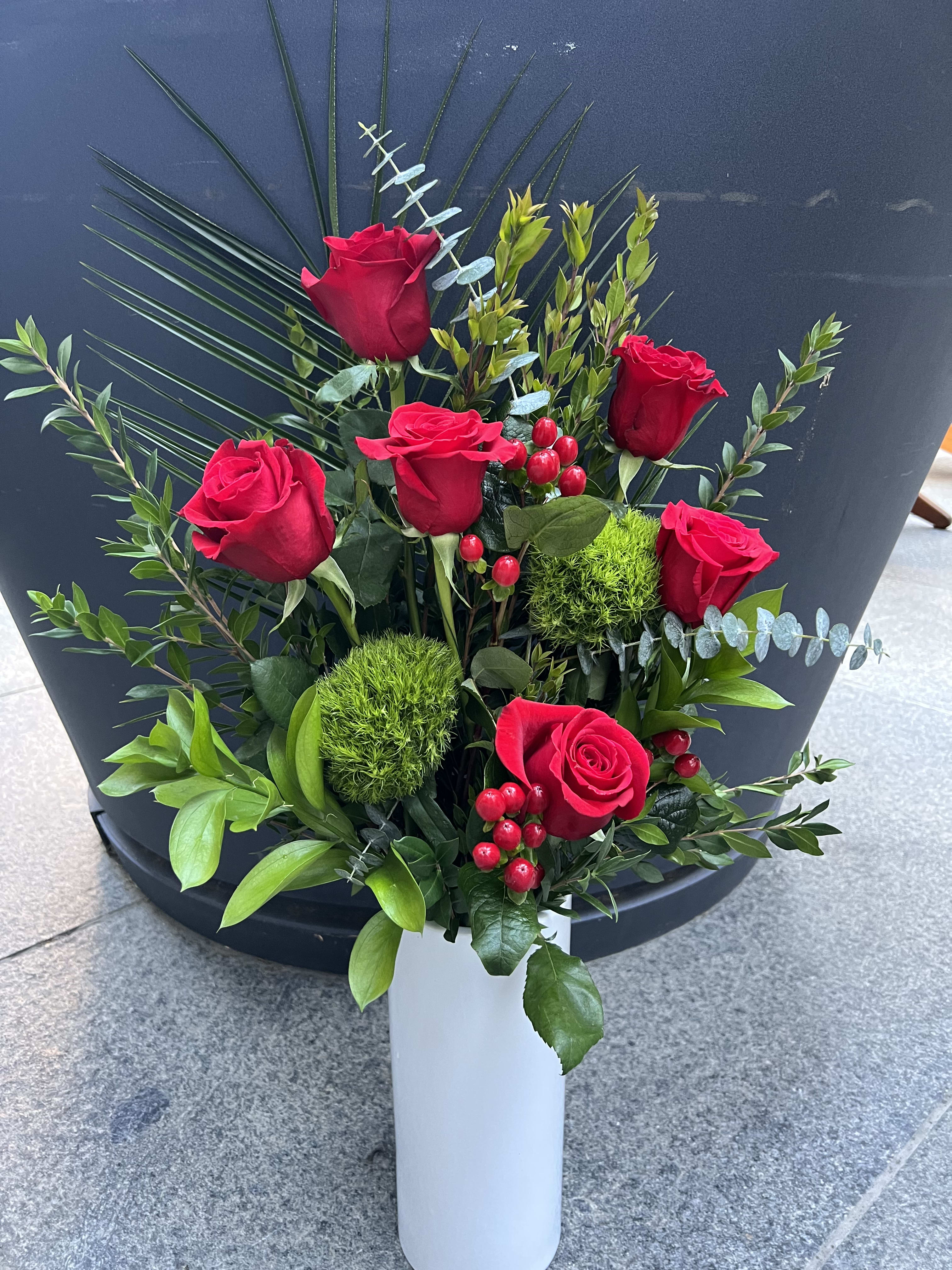 Red roses arranged in a white vase with greenery and red berries