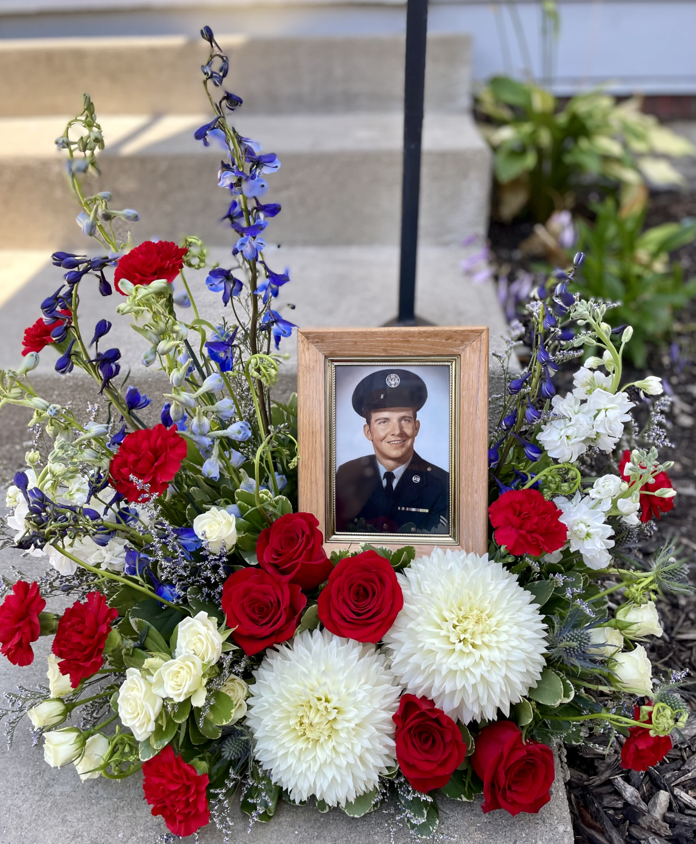 Memorial floral arrangement with a framed portrait, red roses, white blooms, and blue flowers