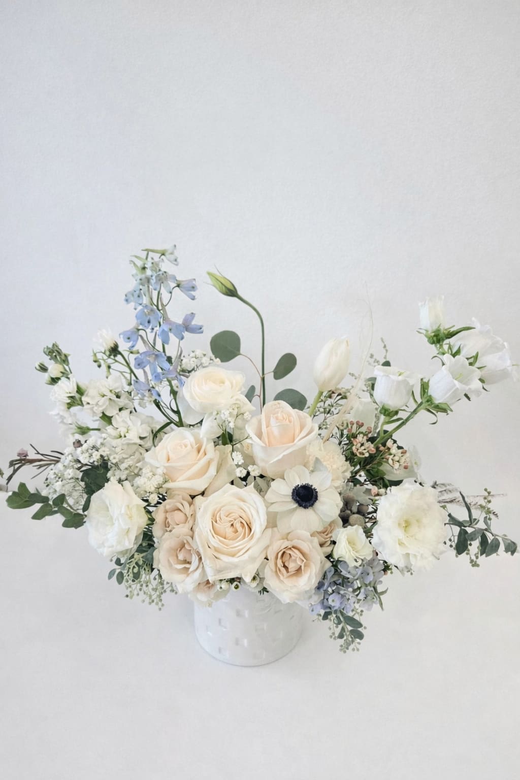 Low arrangement of ivory roses, white blooms, and pale blue flowers in a white ceramic vase