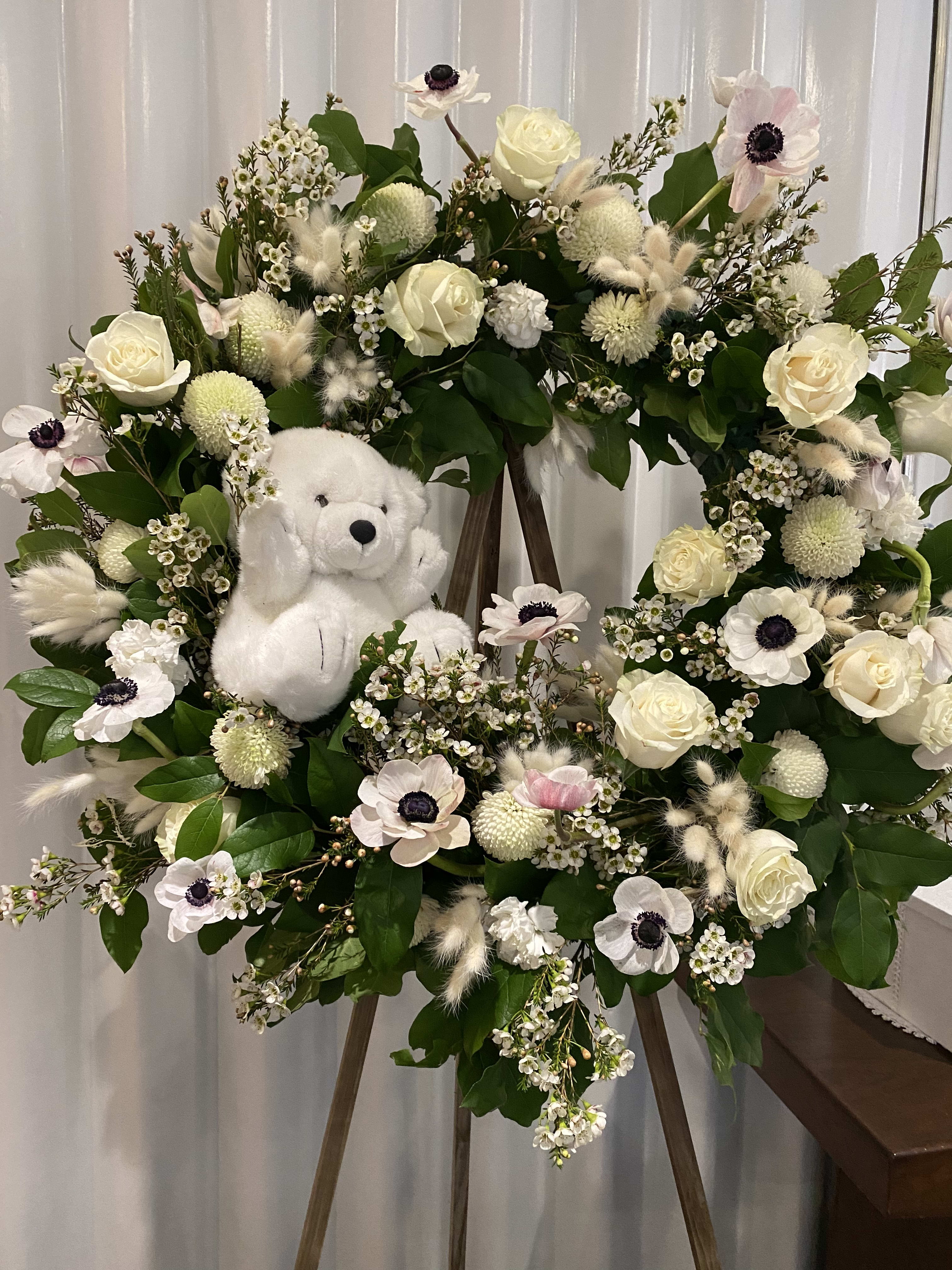 White floral wreath on an easel with a plush teddy bear in the center