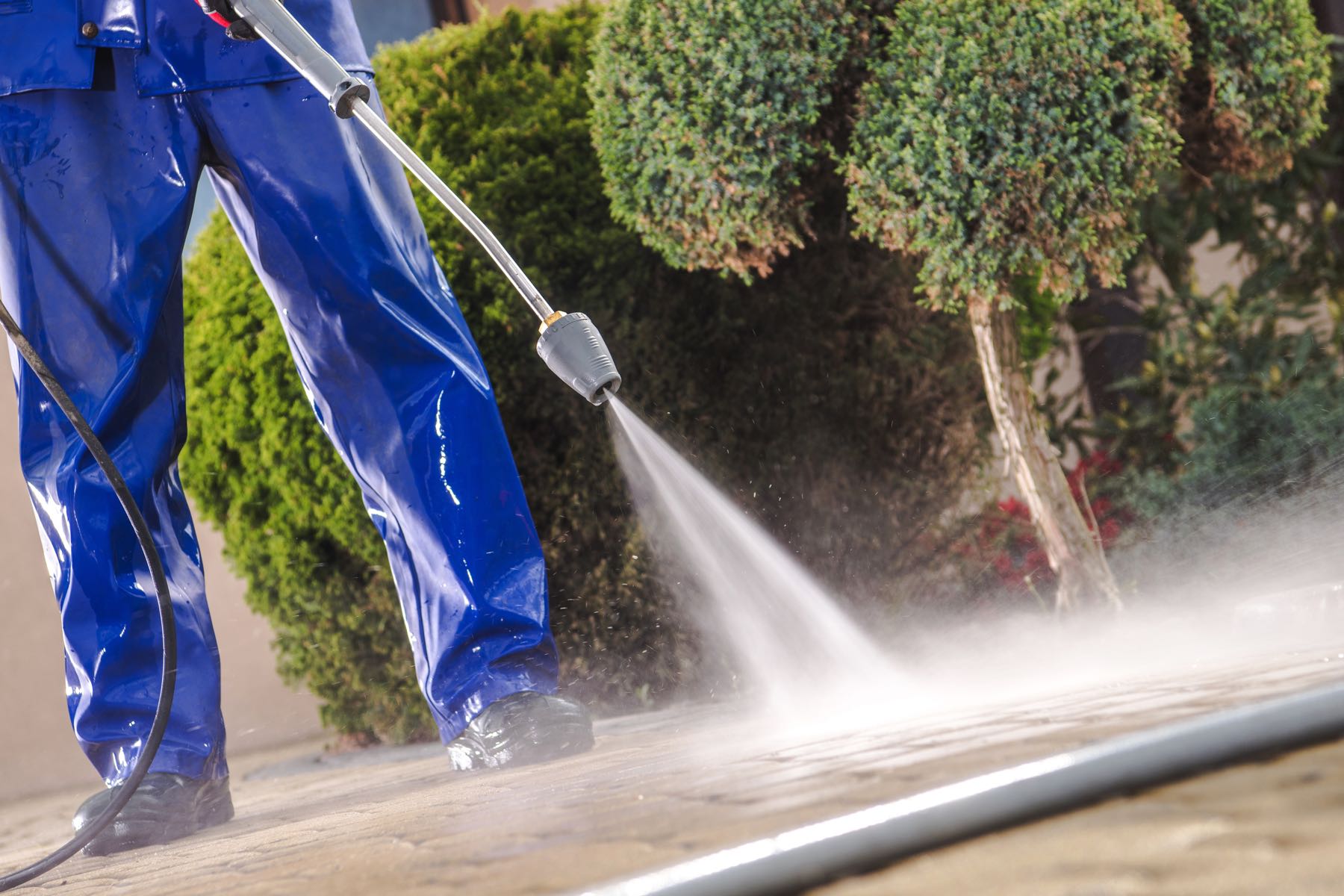 Person in blue protective pants pressure-washing a paved surface outdoors