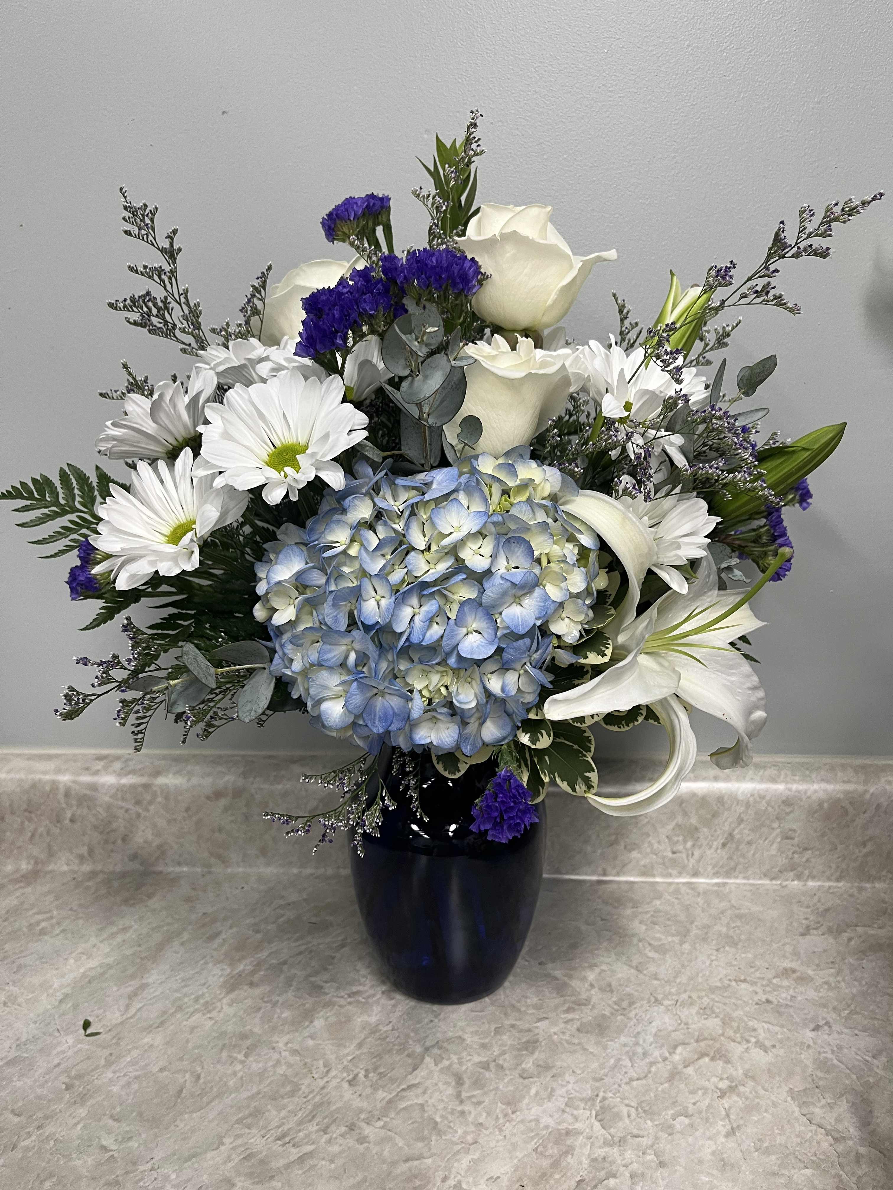 Blue hydrangea bouquet with white roses, lilies, and daisies in a dark vase