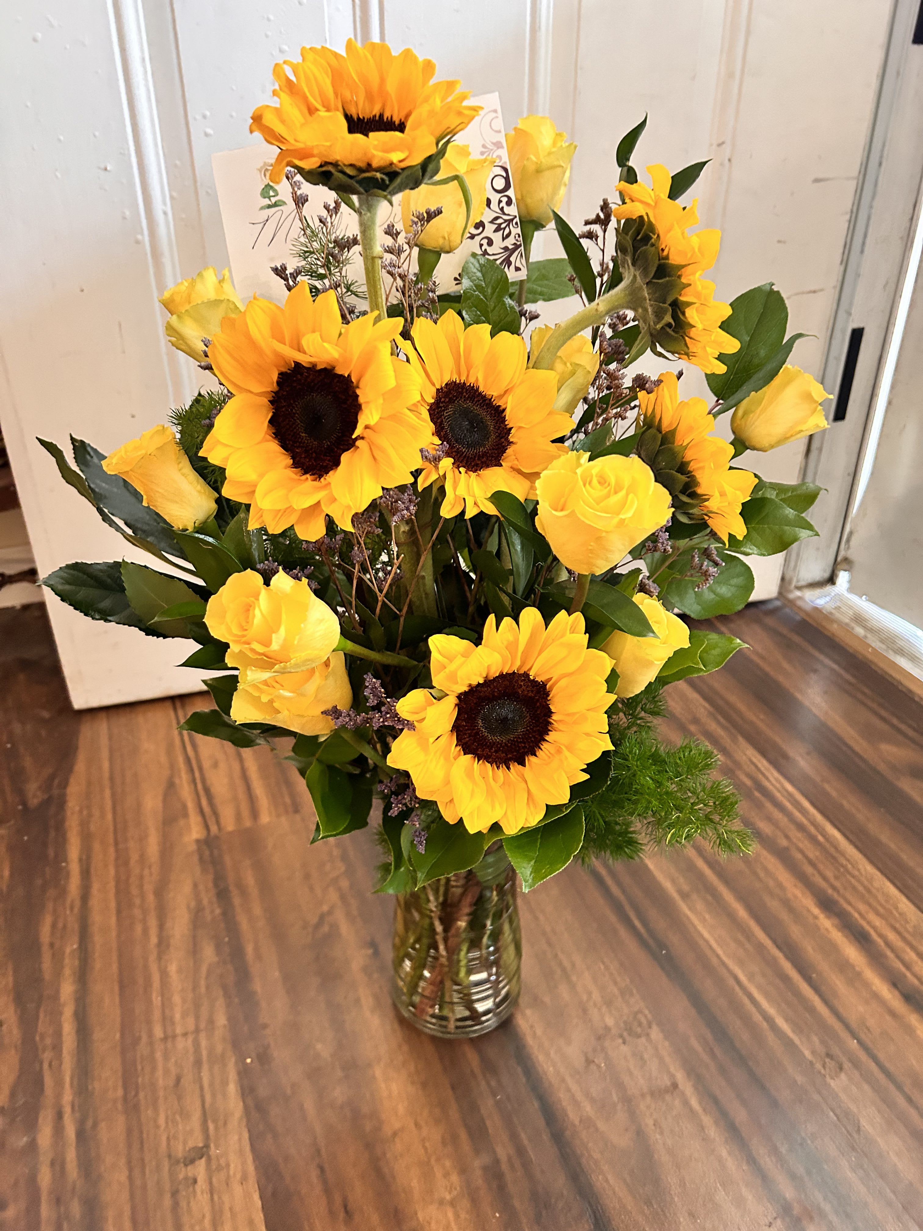 Tall vase arrangement of yellow sunflowers and yellow roses with a small card.