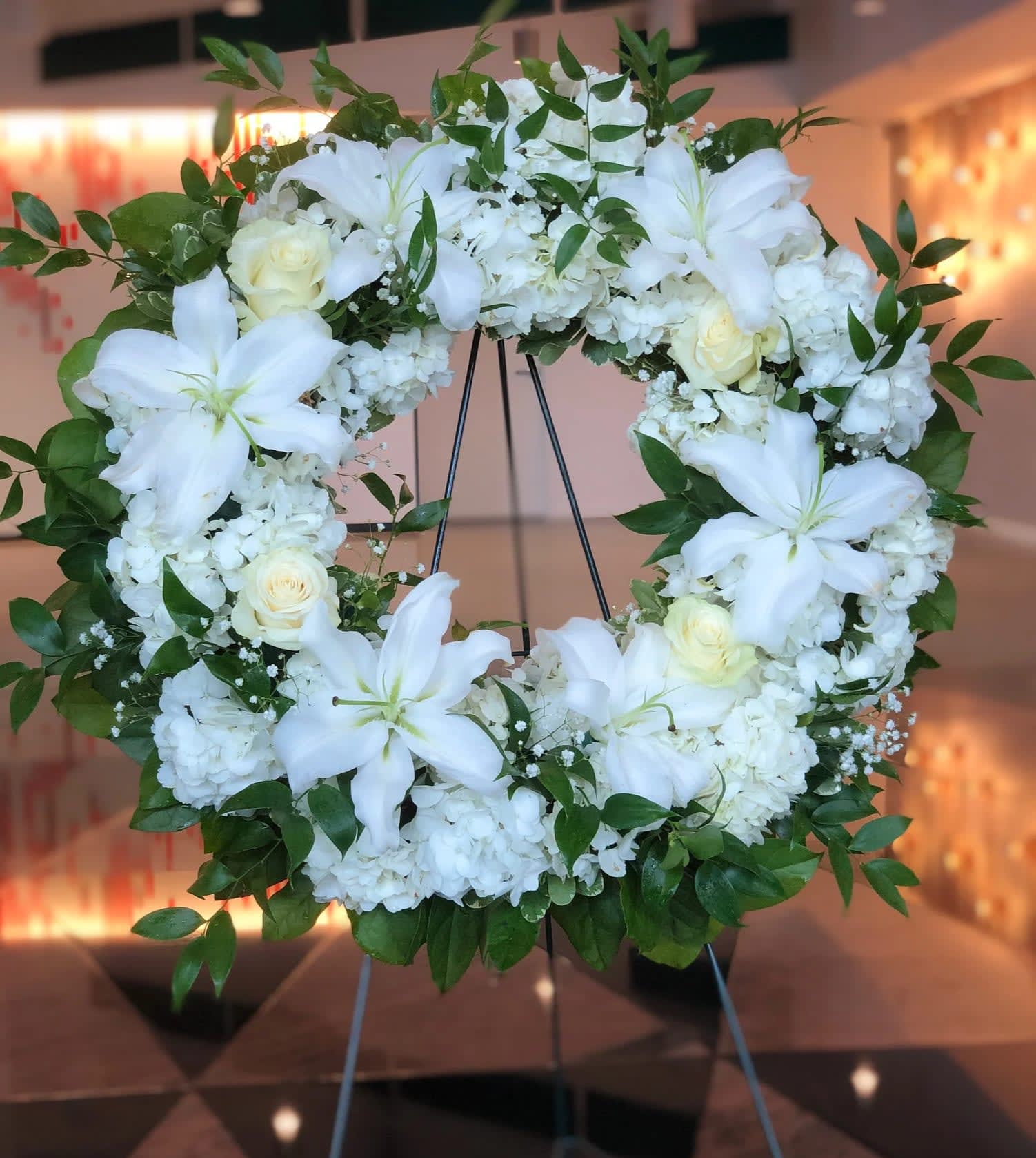 White floral wreath on a stand with lilies, roses, and hydrangeas
