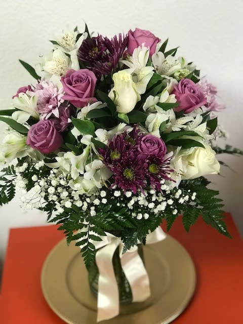 Round arrangement of purple and white flowers in a clear vase with white ribbon on a gold plate