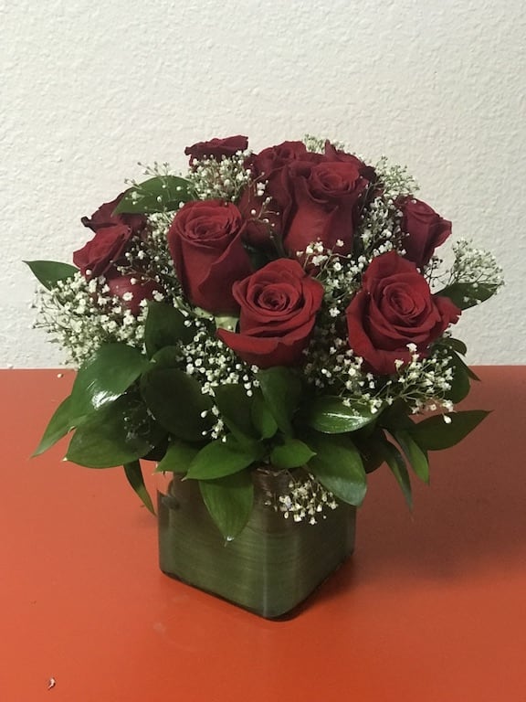 Compact arrangement of red roses with white filler flowers in a green cube vase on an orange surface