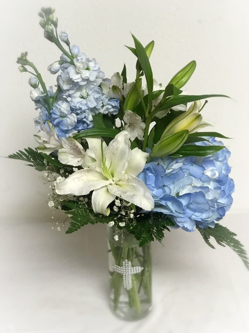 Tall glass vase with blue hydrangeas, white lilies, and other white flowers with a pearl cross on the vase