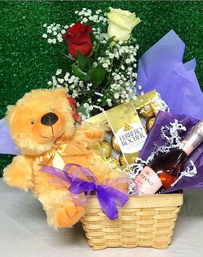 Gift basket with teddy bear, red and white roses, chocolates, and a pink sparkling drink wrapped with purple ribbon