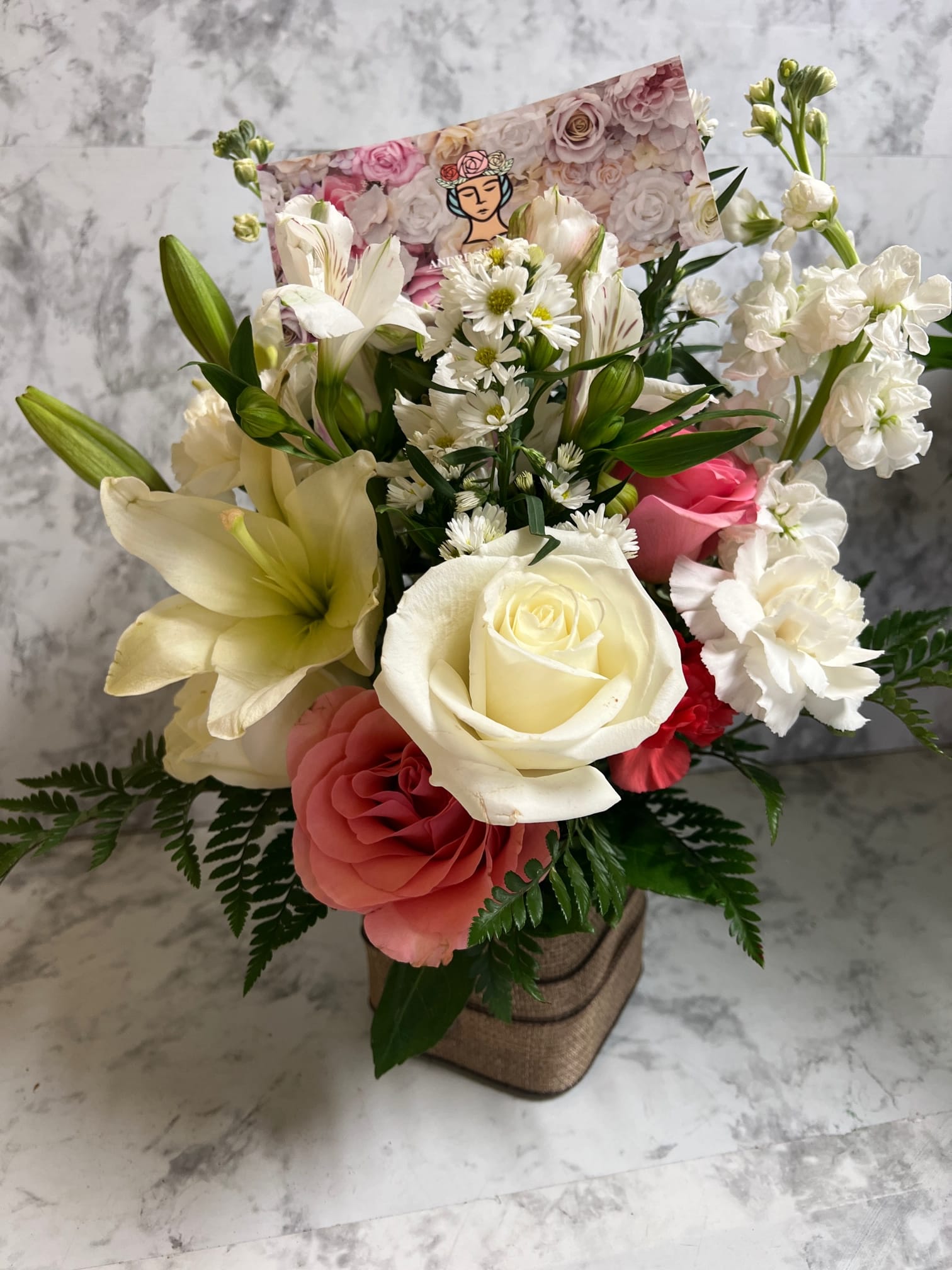 Mixed bouquet of white lilies, roses, and pink blooms in a burlap-wrapped cube vase with a floral card.