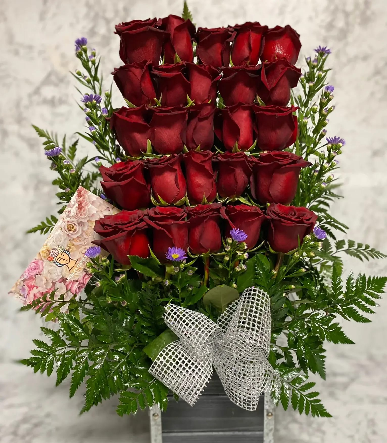 Tall arrangement of tiered red roses with small purple asters in a metal container with a silver bow