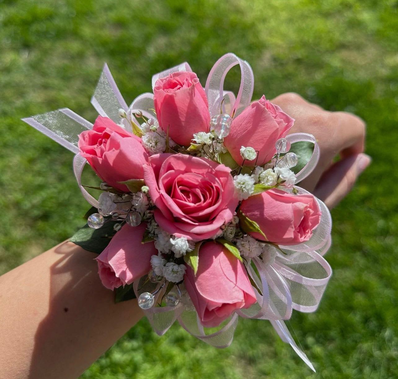Pink rose wrist corsage with white filler flowers, ribbon loops, and bead accents on a person's wrist