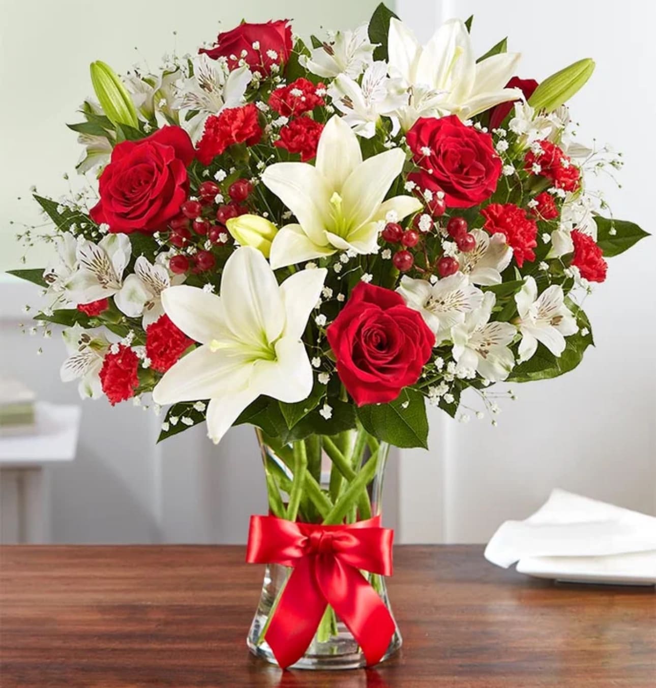 Arrangement of red roses, red carnations, and white lilies in a clear glass vase with a red ribbon bow