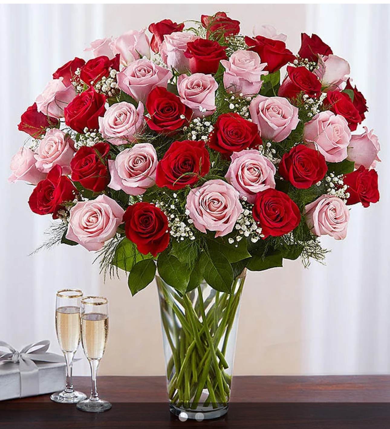 Tall clear vase of red and pink roses with baby's breath, next to two champagne glasses and a silver gift box.