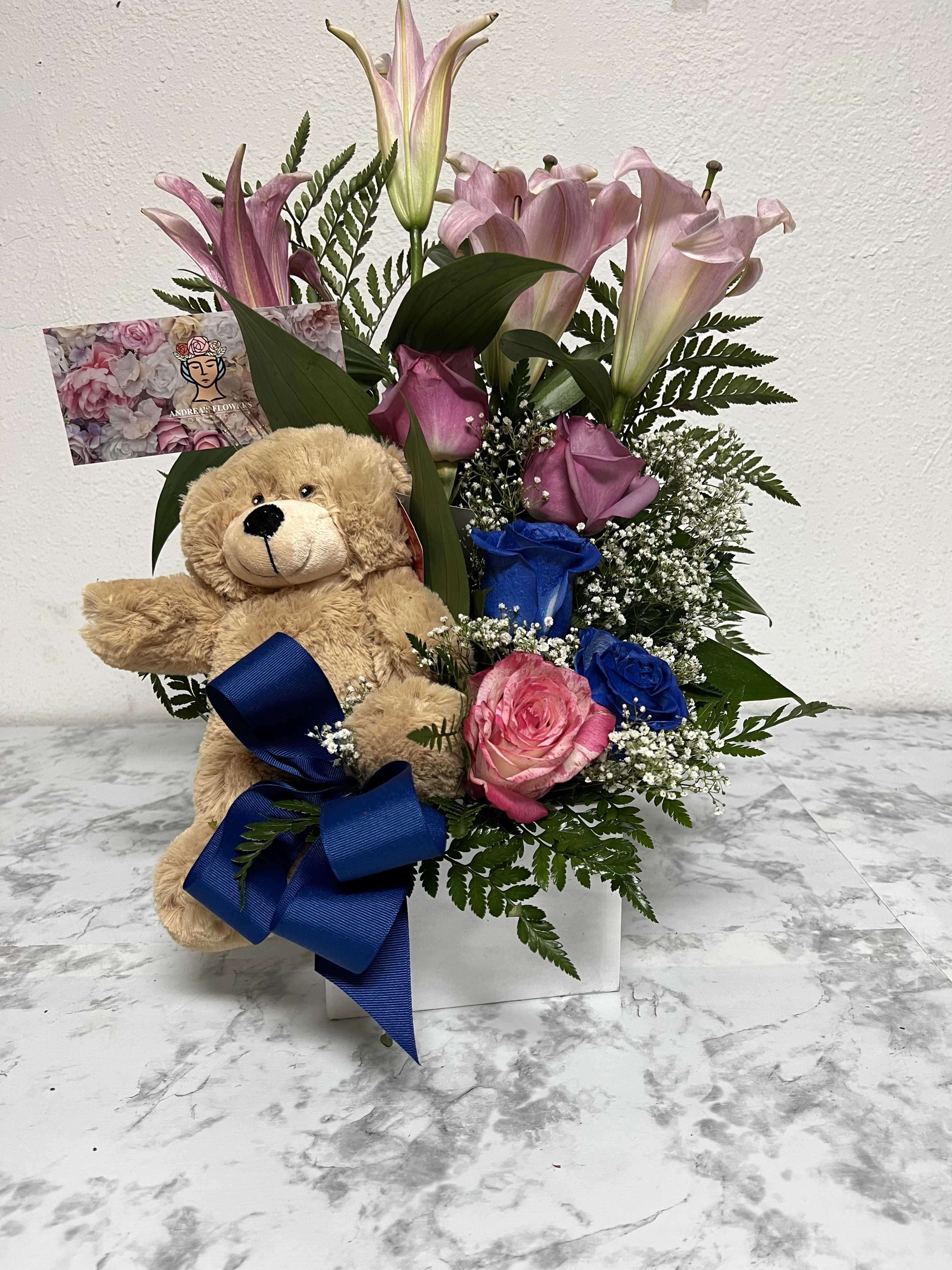 Pink lilies and roses with blue roses in a white container alongside a teddy bear and blue bow.