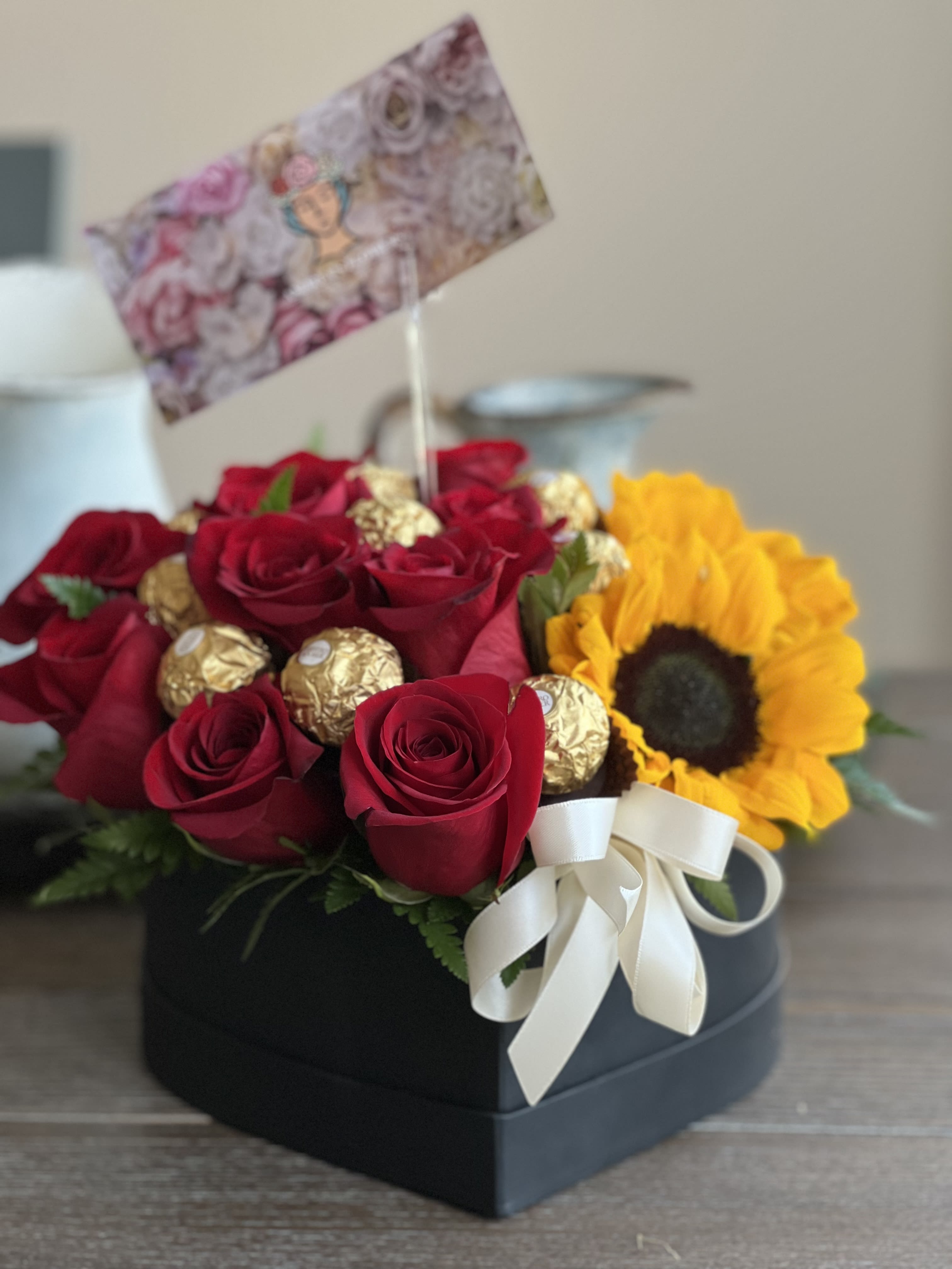 Red roses and a sunflower in a black box with gold chocolates and a white ribbon