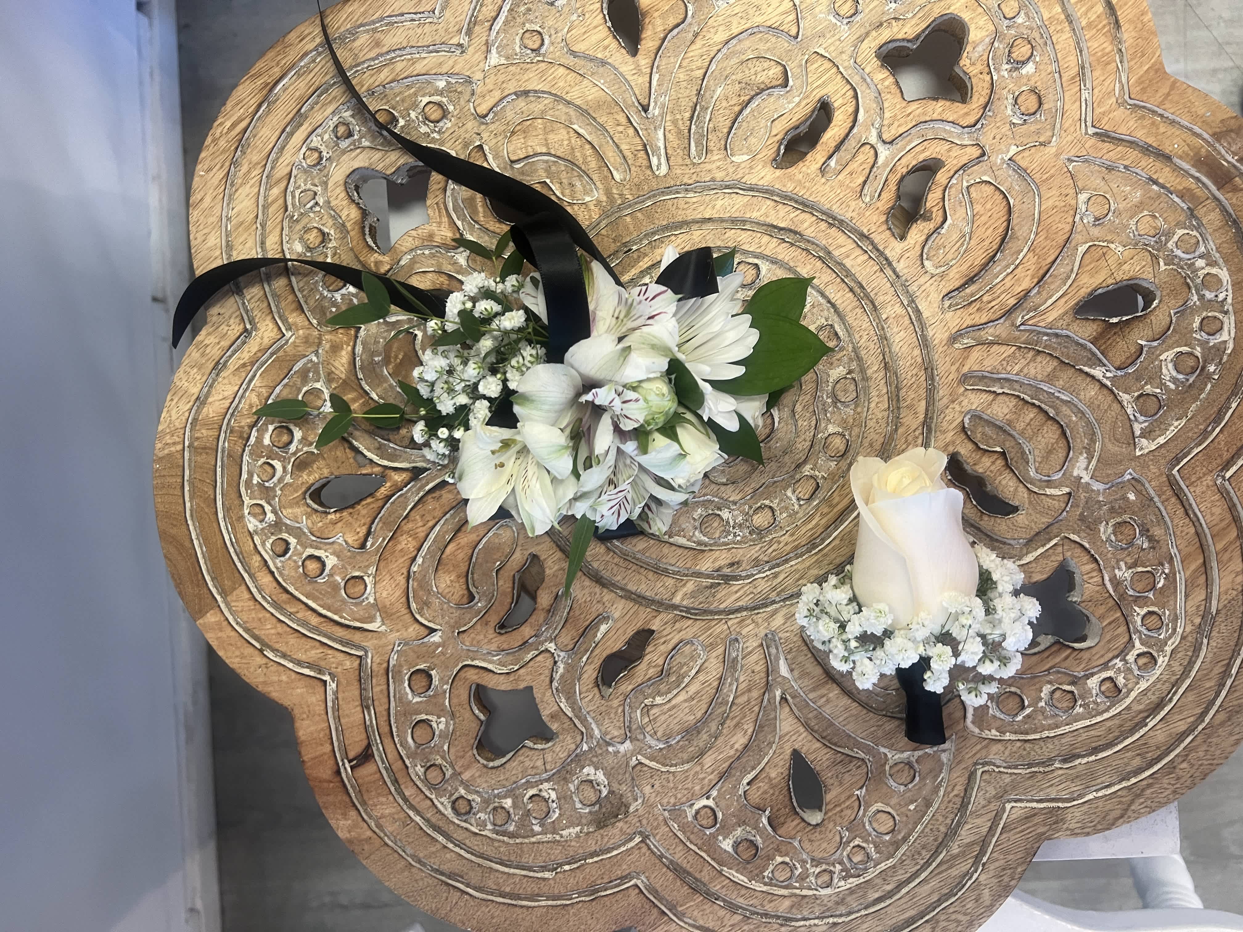 White floral corsage with black ribbon and matching white rose boutonniere on carved wooden table